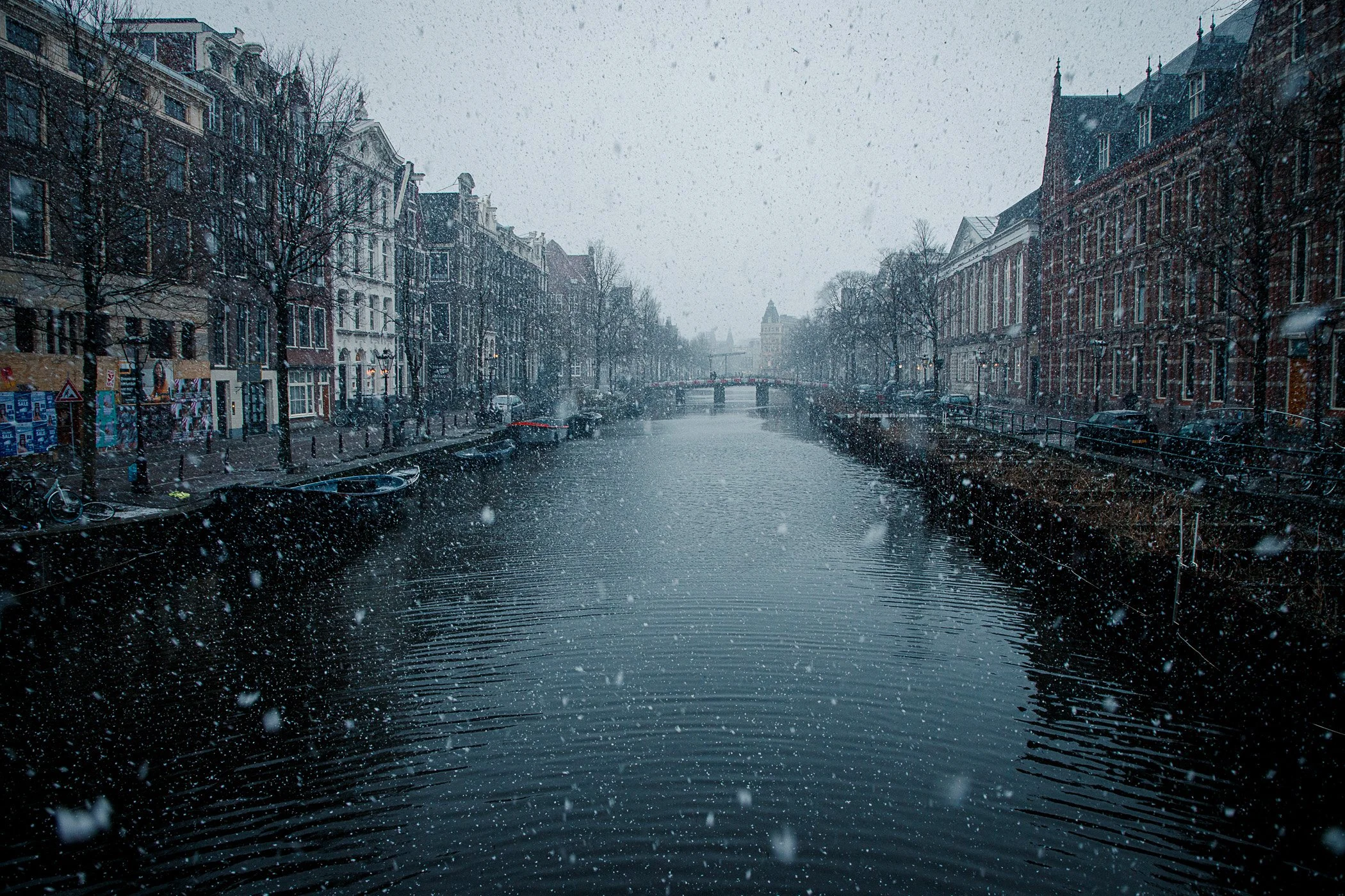 A canal with buildings lining both sides, snow falling heavily, and a bridge in the distance.