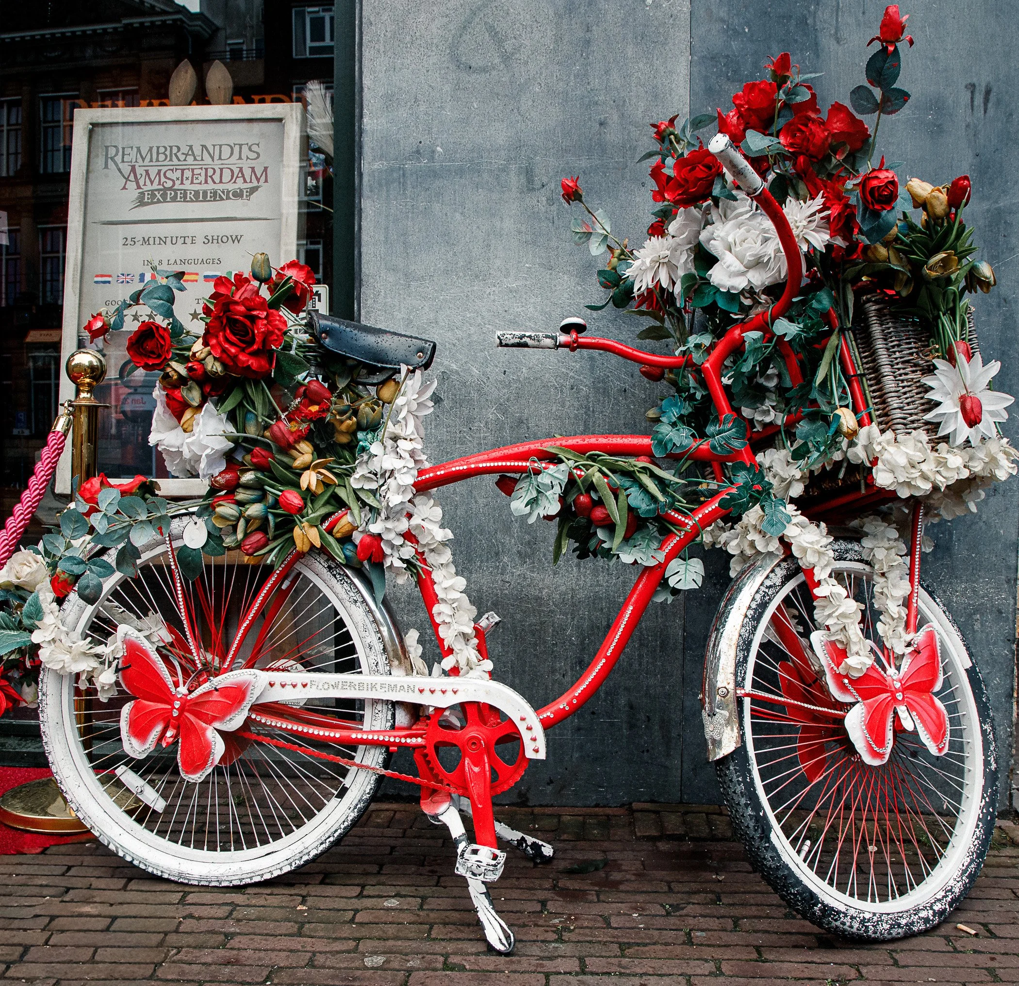 A red bicycle decorated with red and white flowers, leaves, and large butterfly ornaments on the wheels and chain guard, parked outside near a gray wall and a signboard.