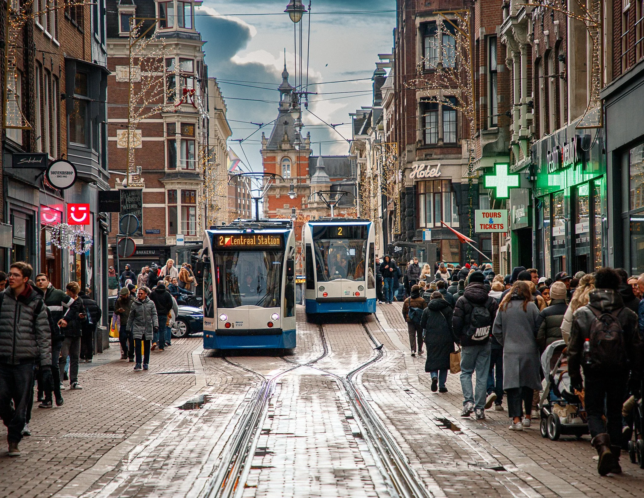A busy city street with two modern trams traveling on tracks, surrounded by pedestrians walking on cobblestone pavement, with shops, a pharmacy, and historic brick buildings lining the street. There are holiday decorations and a mix of people in wint