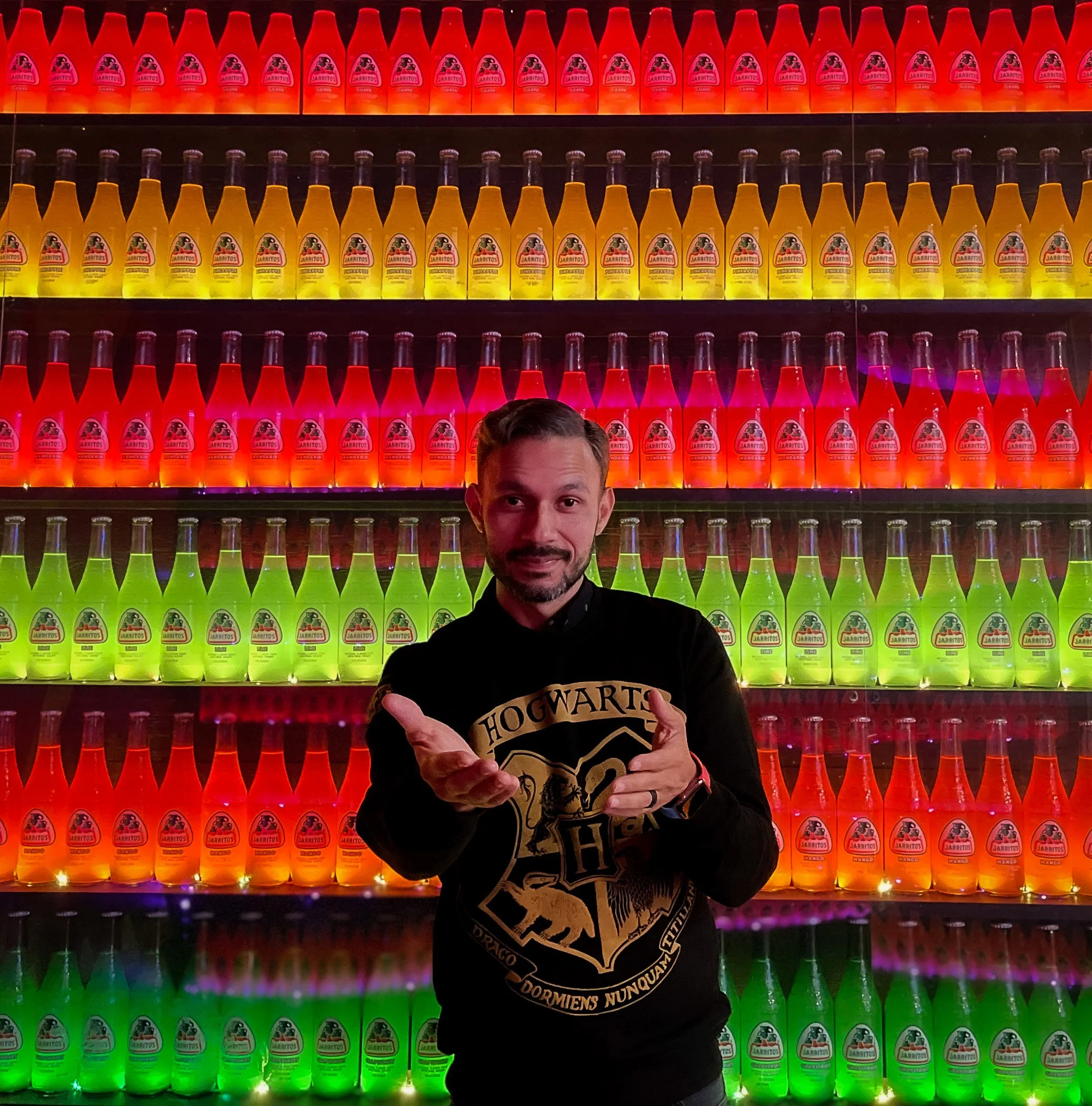 A man standing in front of a colorful display of bottles illuminated with multicolored lights, wearing a black hoodie with the Harry Potter crest.