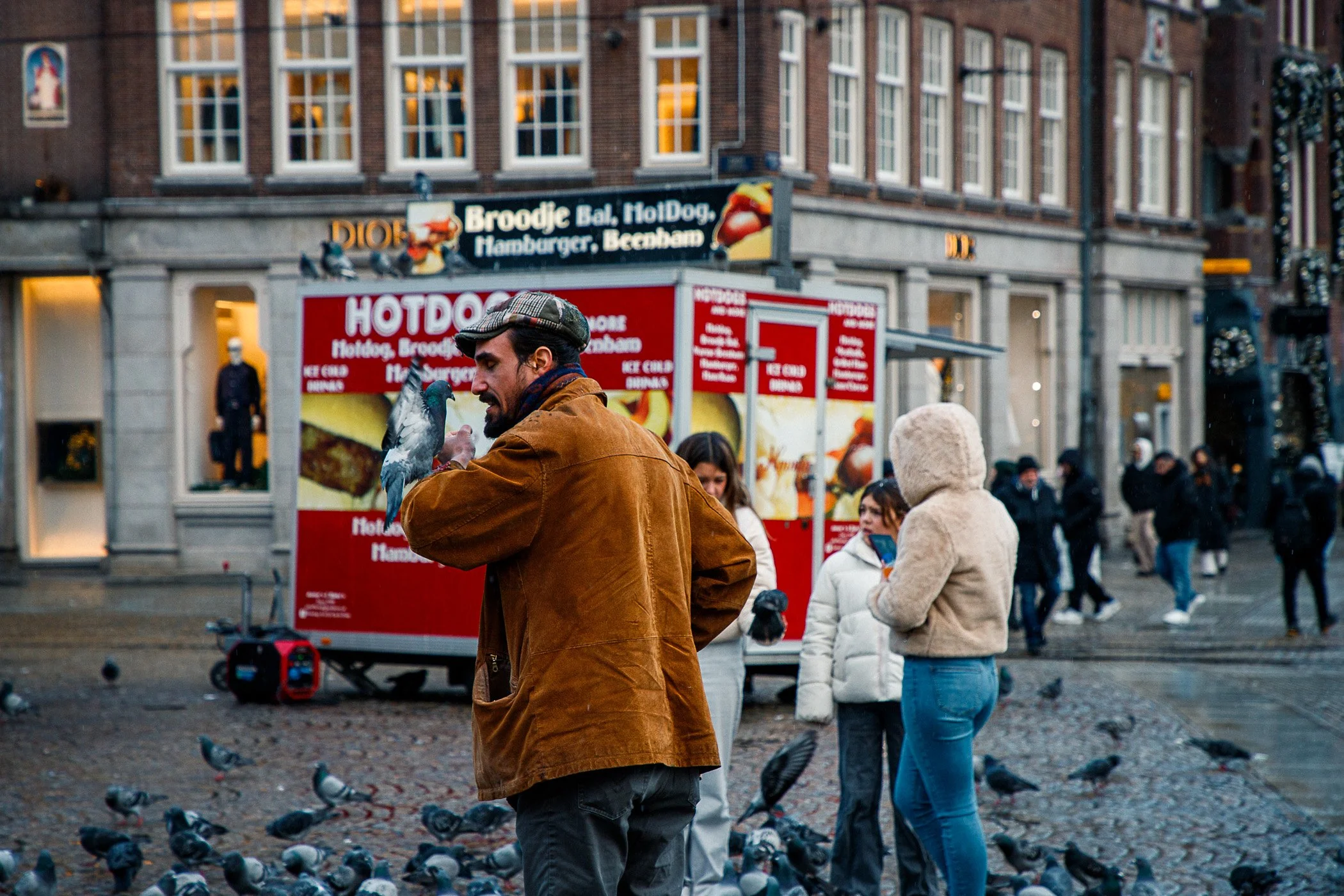 A man feeding pigeons in a busy city square with a street food stand and several pedestrians in the background.