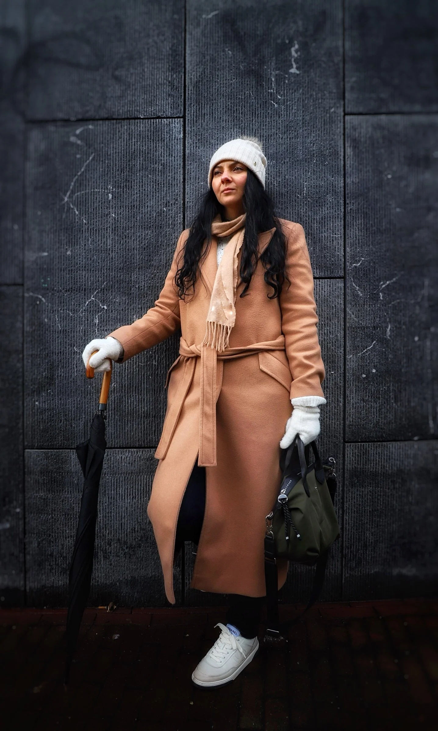 A woman with long dark hair standing against a dark textured wall, wearing a beige coat, white beanie, beige scarf, white gloves, white sneakers, holding an umbrella in her right hand, and a black and green bag in her left hand.