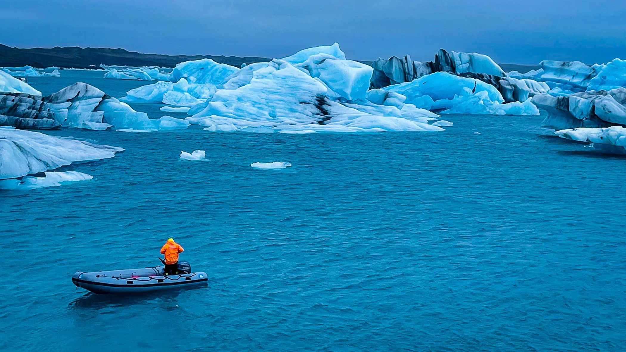 A person in an orange jacket standing on an inflatable boat in icy waters surrounded by large icebergs.