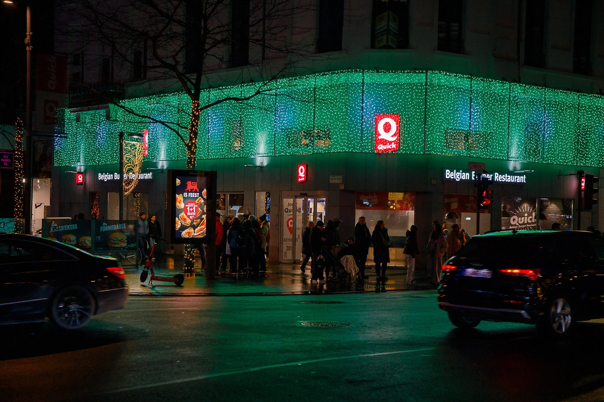 Night view of a busy city street corner with a brightly lit restaurant sign that says 'Quick' and surrounded by Christmas decorations and string lights. Pedestrians and cars are visible.