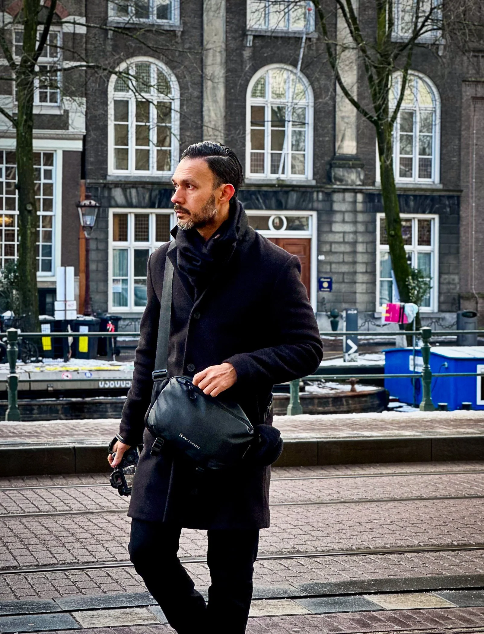 A man with dark hair and beard, wearing a dark coat and scarf, walks along a city street with tram tracks. Behind him is a canal with a boat, and buildings with large arched windows and trees.