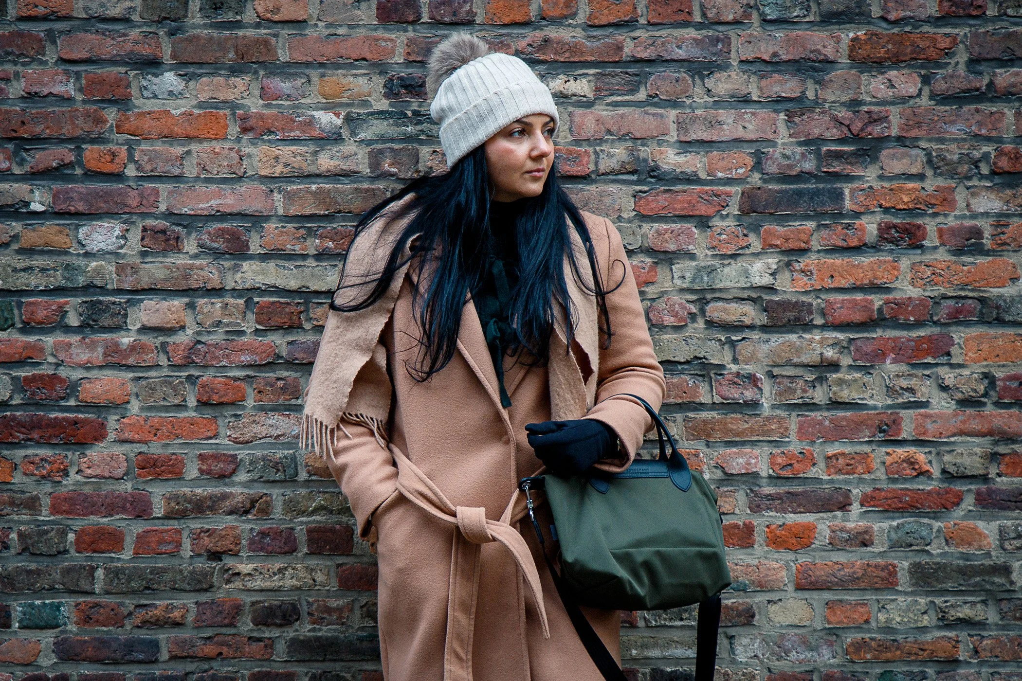 A woman with long dark hair, wearing a beige coat, black gloves, and a beige knit beanie with a pom-pom, standing against a brick wall. She is holding a green bag and looking to her right.