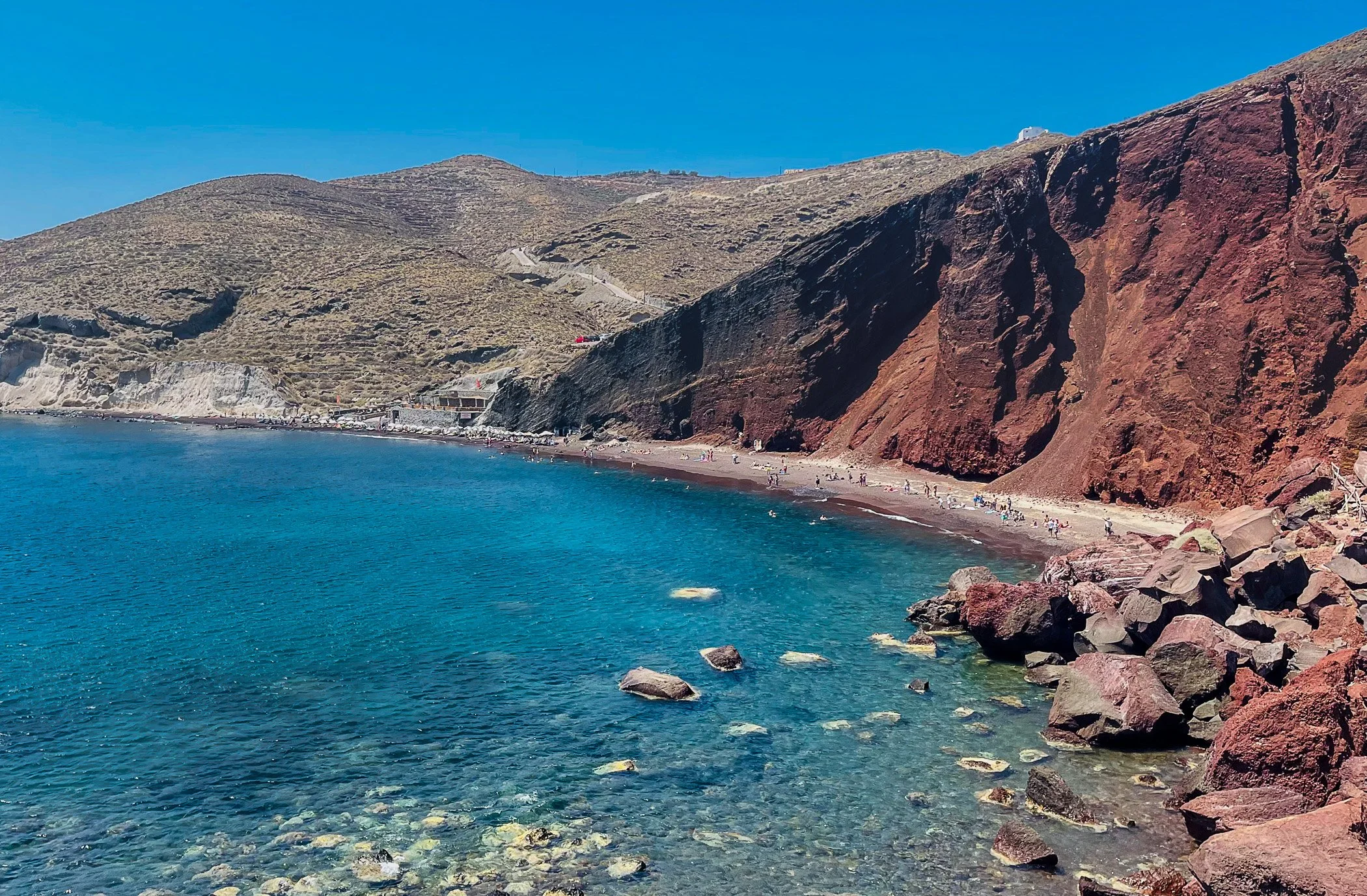 Coastal landscape featuring a sandy beach with people, brown and red cliffs, and a clear blue sky.