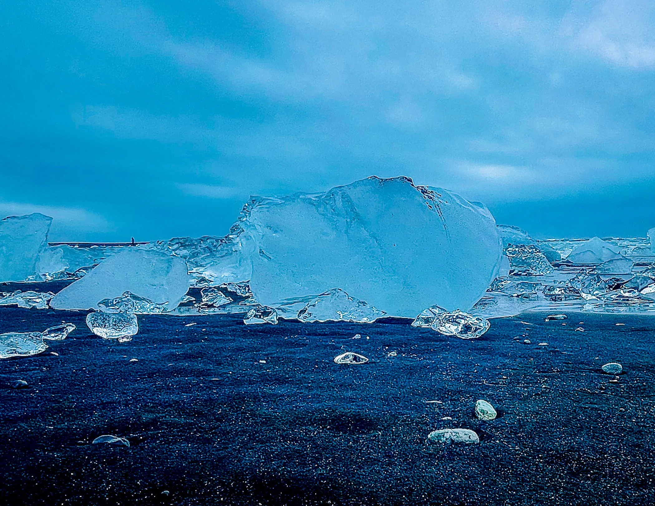 Icebergs and ice fragments scattered on a dark volcanic beach with a cloudy sky in the background.