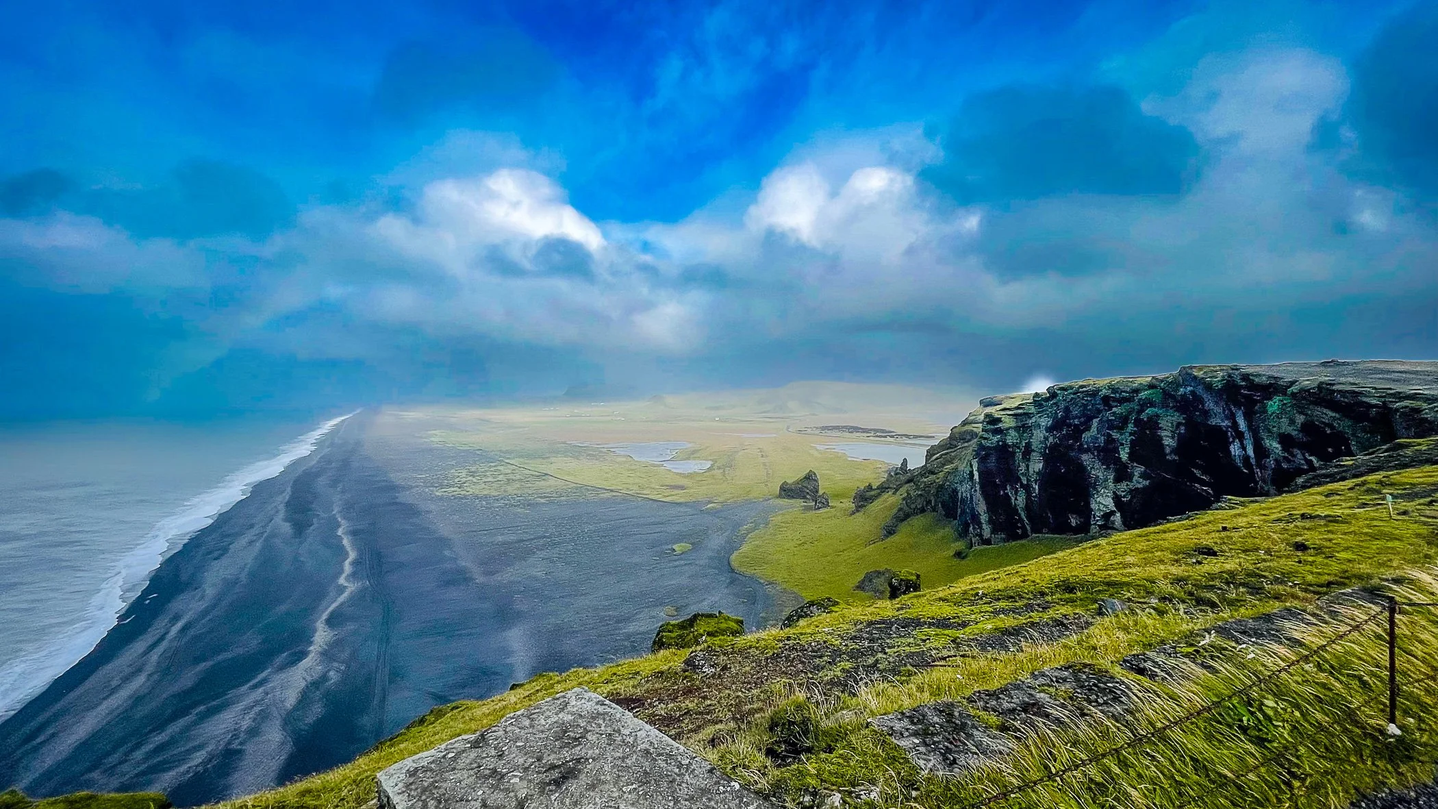 Coastal landscape with a cliff on the right, a black sand beach on the left, grassy hills in the background, and a cloudy sky