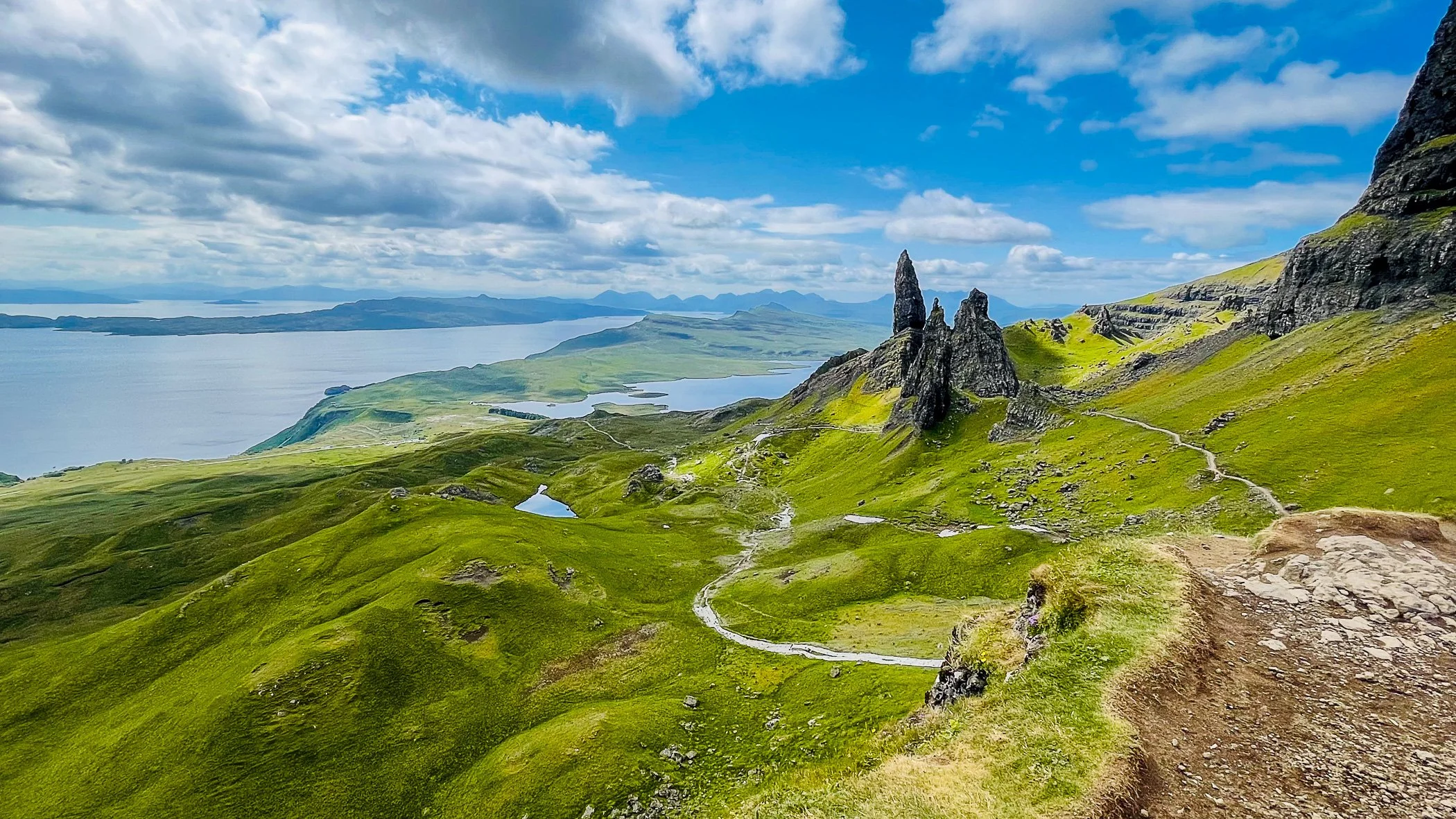 Vivid landscape of green rolling hills with rocky formations and a winding dirt trail, overlooking a large body of water under a partly cloudy sky.