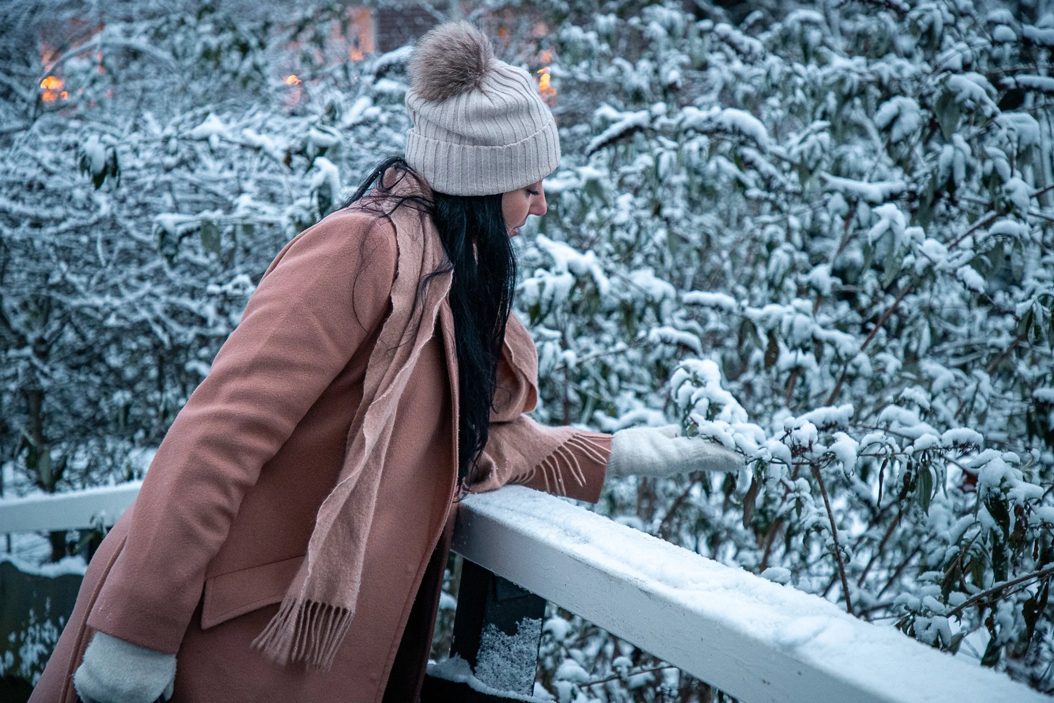 Woman in a beige coat and white knit hat touches snow-covered bushes on a winter day.