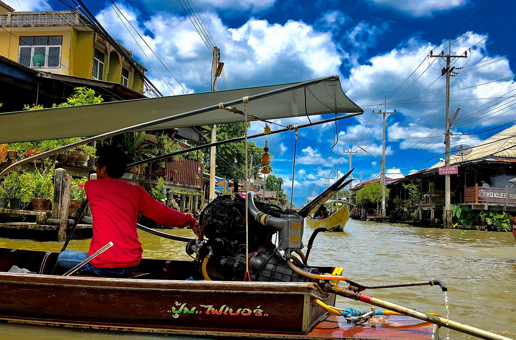 A boat navigating a canal in a town with colorful buildings, power lines, and a lively sky with clouds.