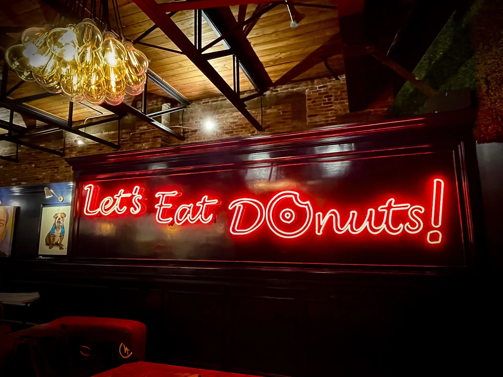 Neon sign reading 'Let's Eat DONUTS!' in a restaurant with exposed brick walls, a wooden ceiling, and modern lighting fixtures.