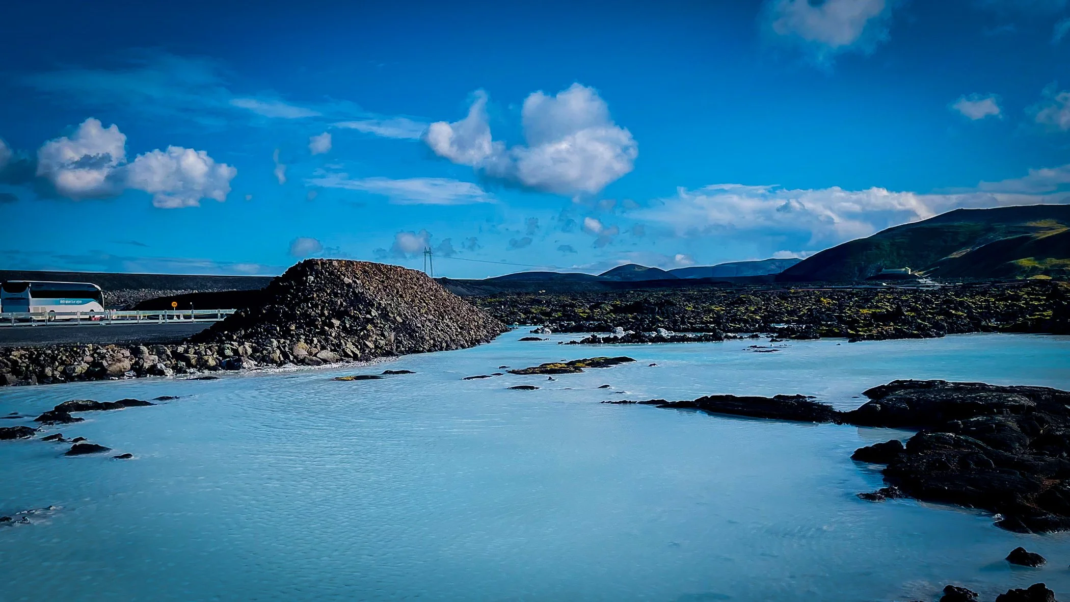 A scenic landscape of a river with clear blue water flowing through a rocky terrain, with a bridge and vehicle on the left, and green mountains in the background under a partly cloudy sky.