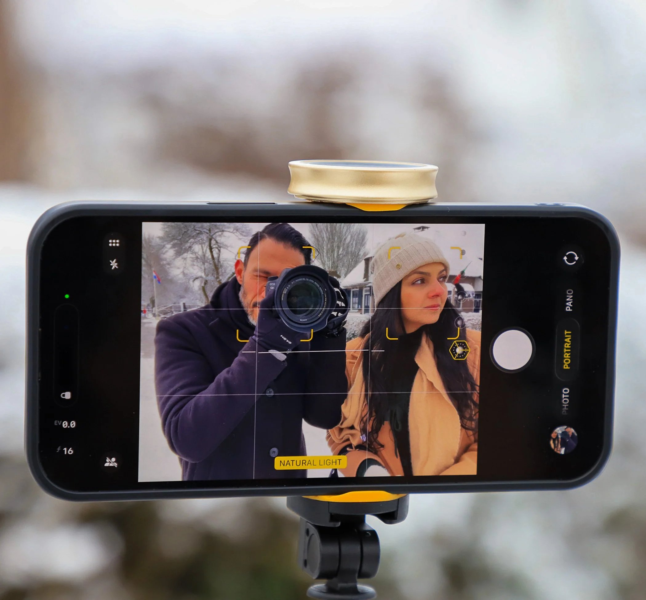 A smartphone camera captures a man and woman in a snowy outdoor setting, with the man holding a camera and the woman wearing a beige hat.