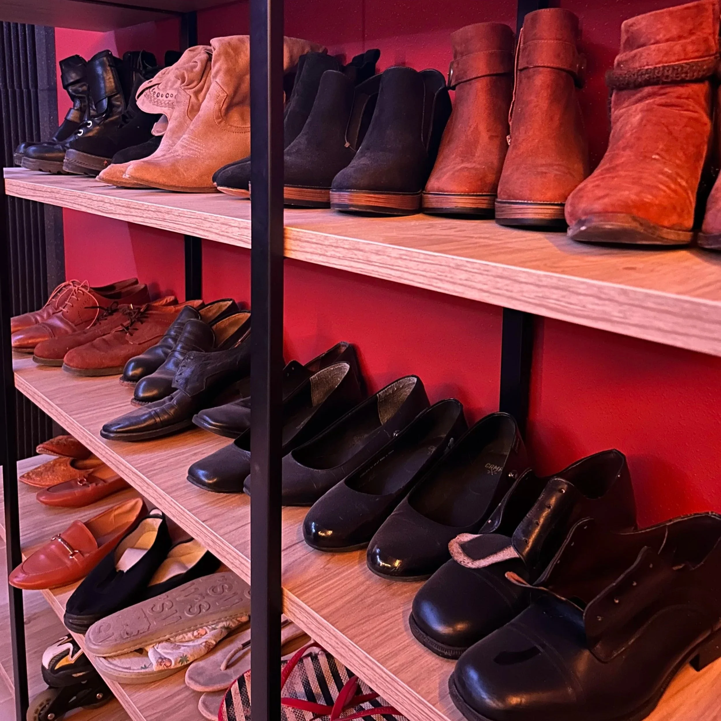 A display shelf with various shoes, including boots, dress shoes, and slippers, arranged on multiple wooden shelves against a red wall.