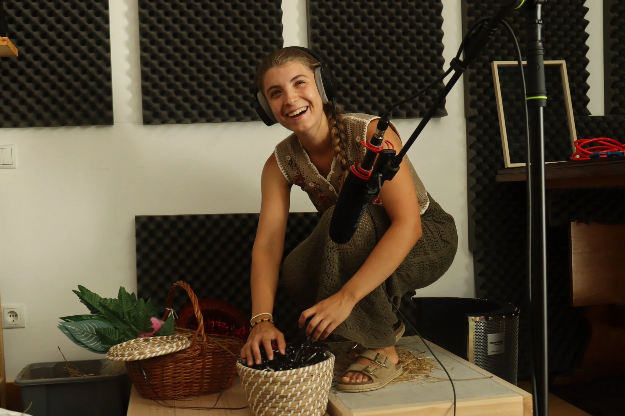 Woman wearing headphones squatting on a platform, smiling, with recording equipment around her, in a soundproofed room.
