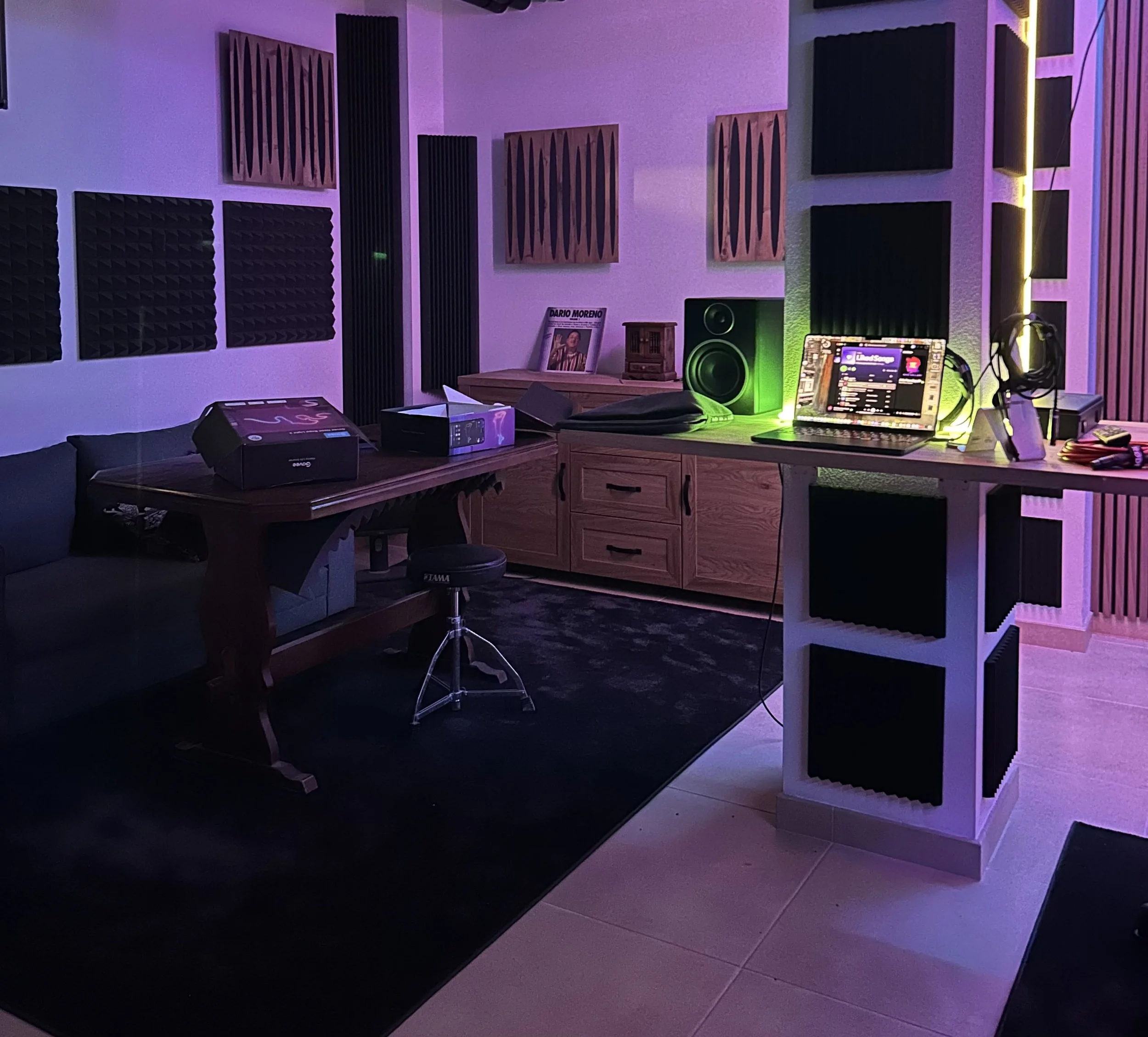 Sound recording studio with soundproofing foam panels, a computer on a stand, studio monitor speakers, a desk with audio equipment, vinyl records, and headphones, illuminated by purple and green lighting.