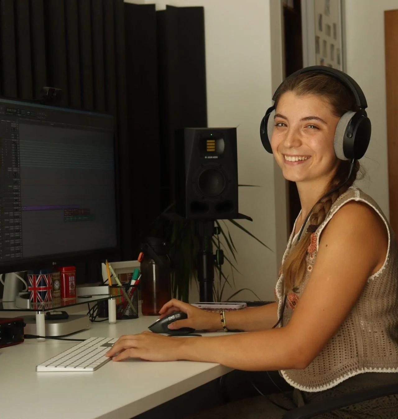 A young woman sitting at a desk with a computer in a music or recording studio, wearing large headphones, smiling at the camera, with sound equipment and supplies on her desk.