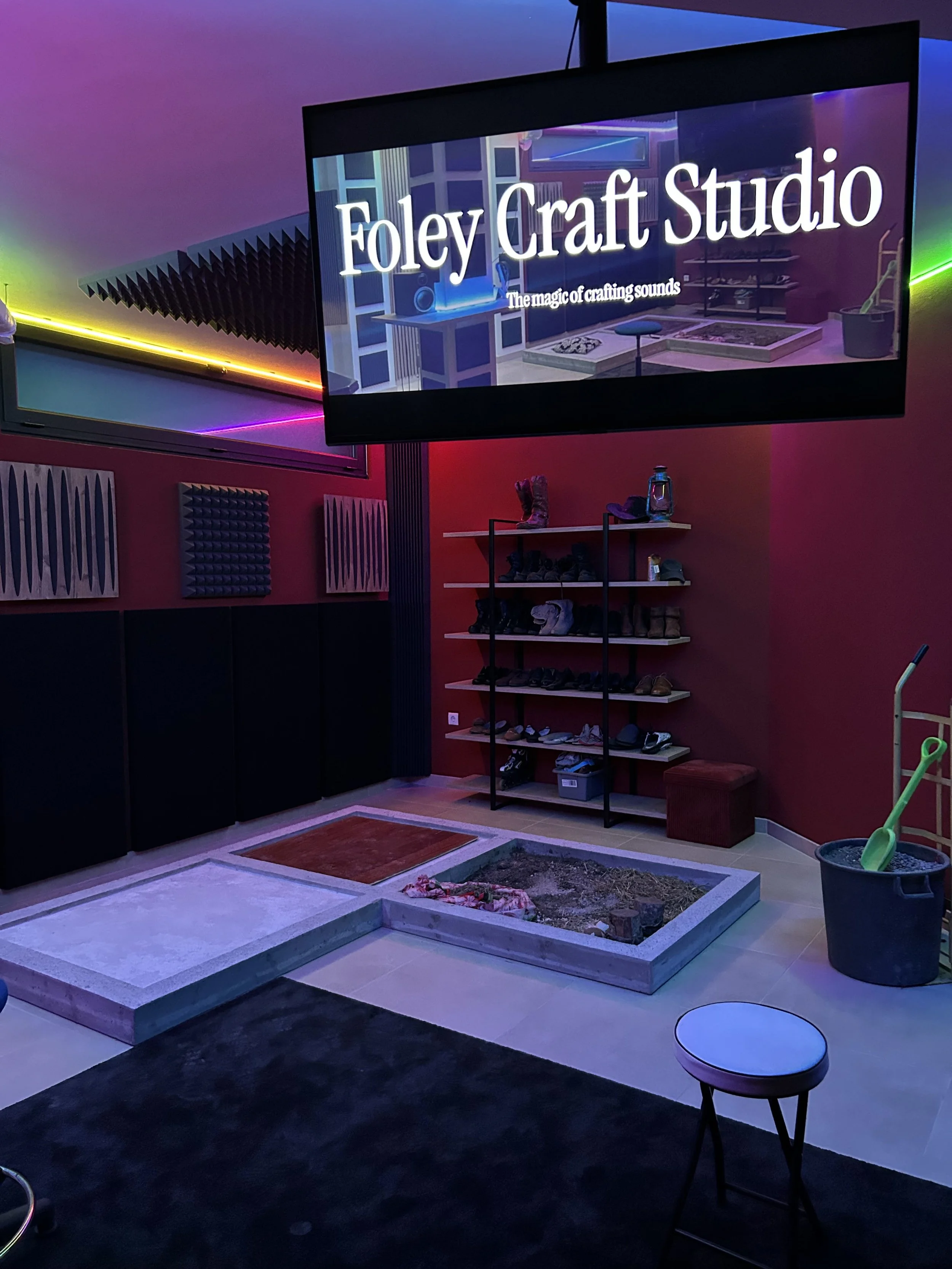 Indoor music or sound studio with a large screen displaying 'Foley Craft Studio' and 'The magic of crafting sounds.' The room is decorated with colorful lighting, acoustic panels, various shoes on a rack, and some gardening tools. There is a small stool and an area with a raised platform, possibly for sound recording.