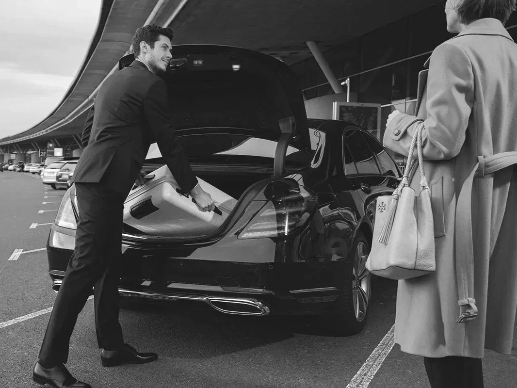 Man loading luggage into trunk of a black car at an airport parking lot, woman standing nearby with a handbag