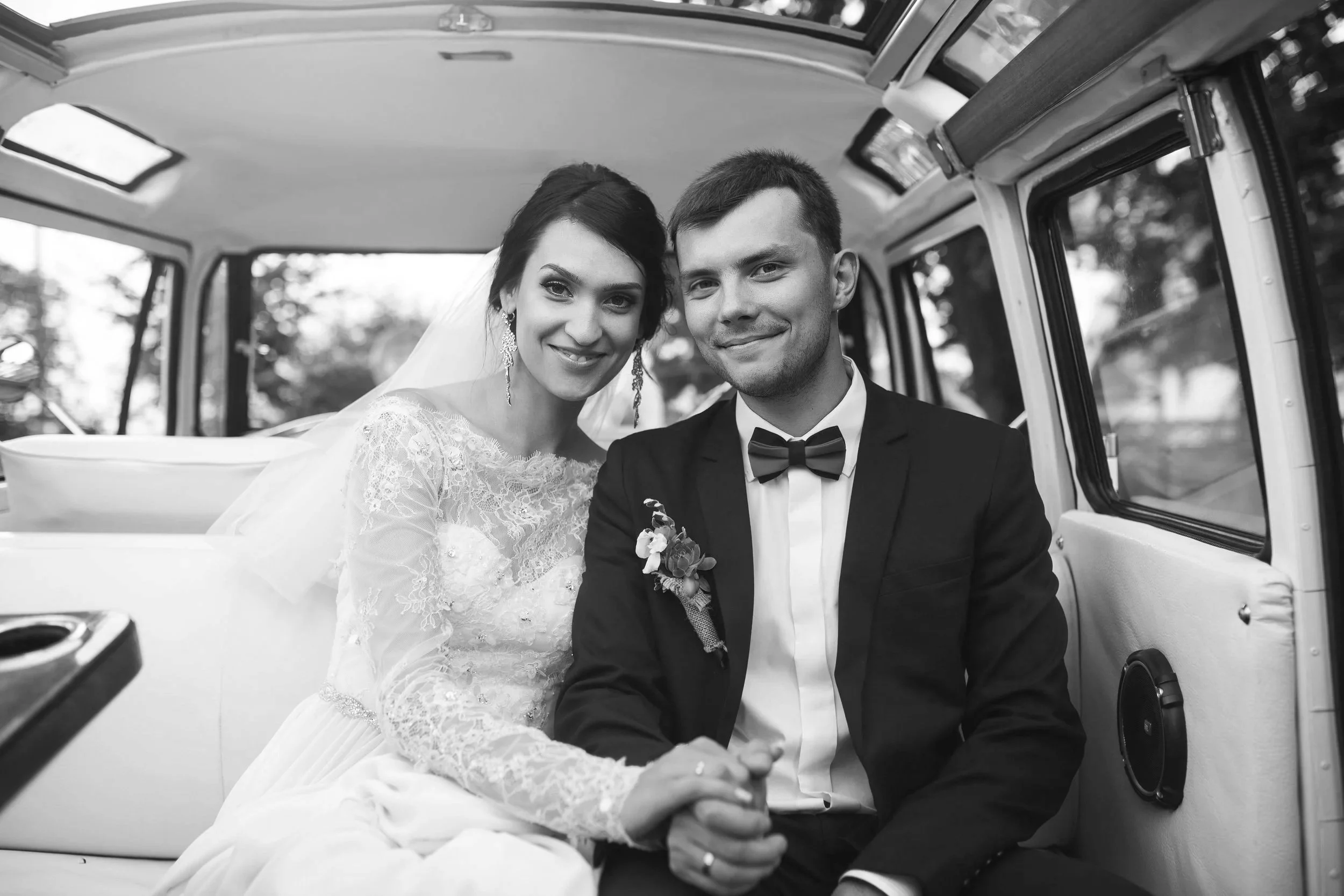 A bride and groom sitting inside a vehicle, smiling and holding hands, during their wedding.