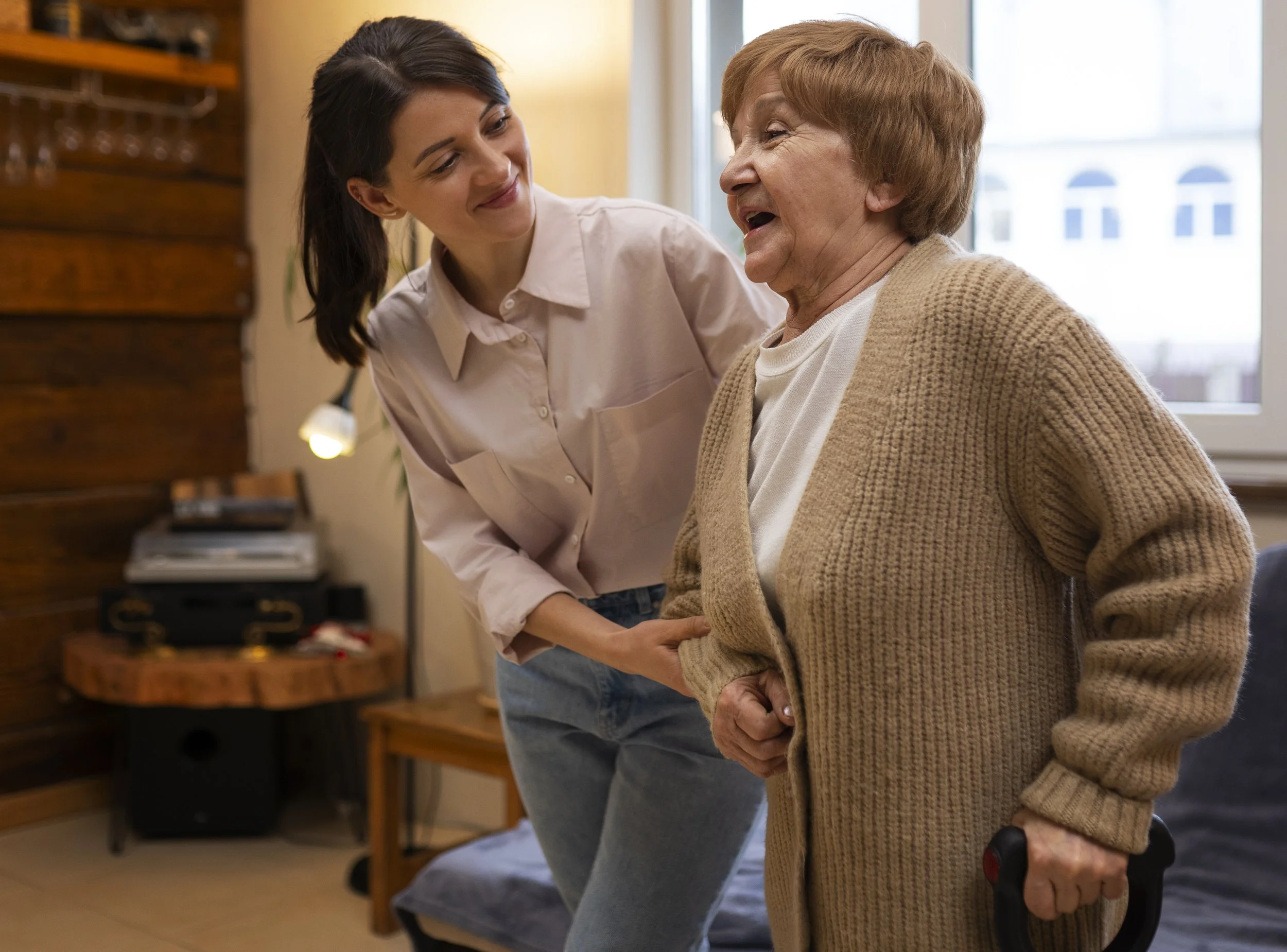 Une jeune femme aide une personne âgée à se lever de sa chaise, dans un intérieur domestique chaleureux.
