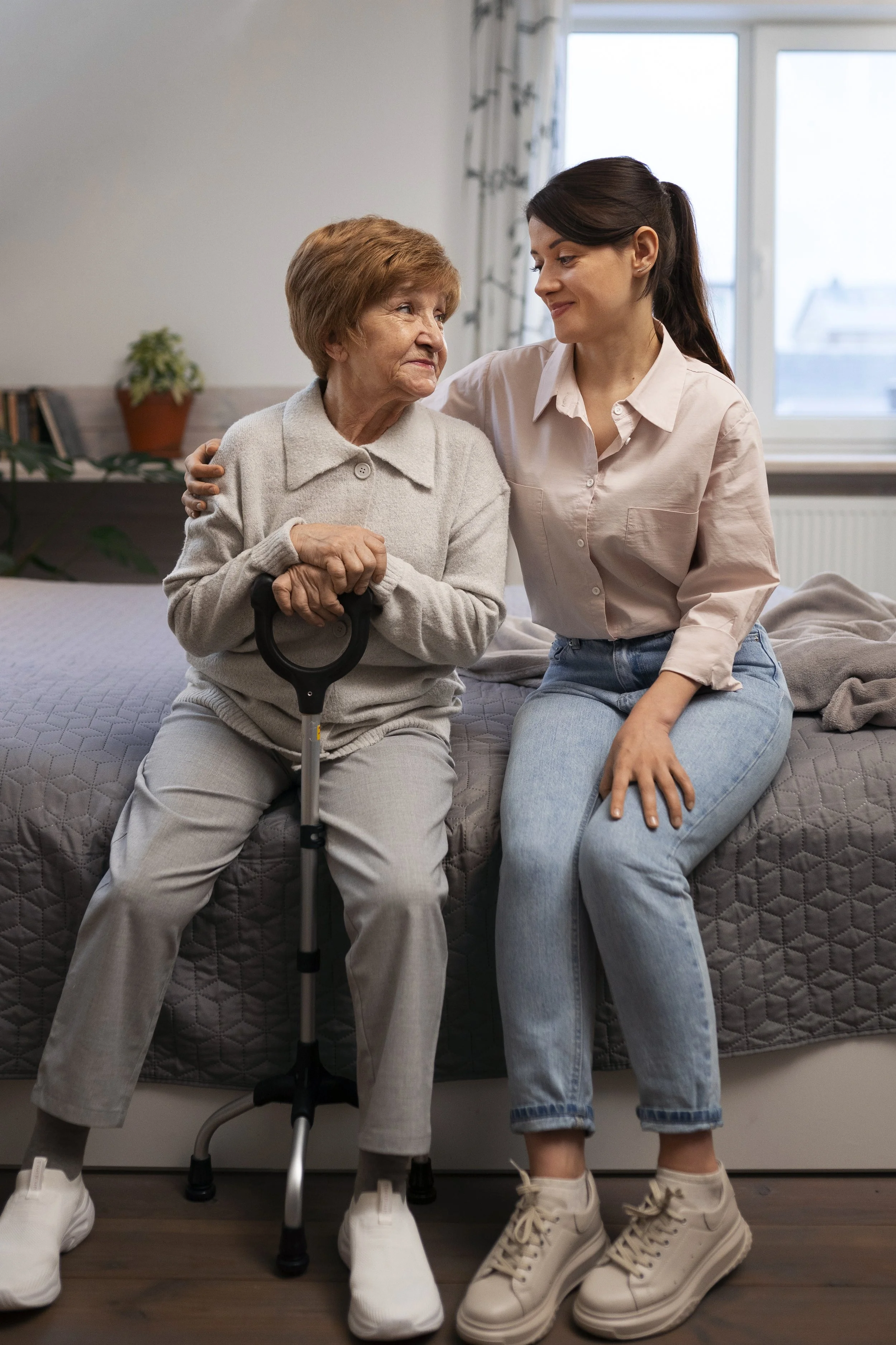 Une jeune femme et une femme âgée s'assoient sur un lit, se regardant avec tendresse, la jeune femme soutenant la personne âgée avec une main sur l'épaule et l'autre sur le genou.