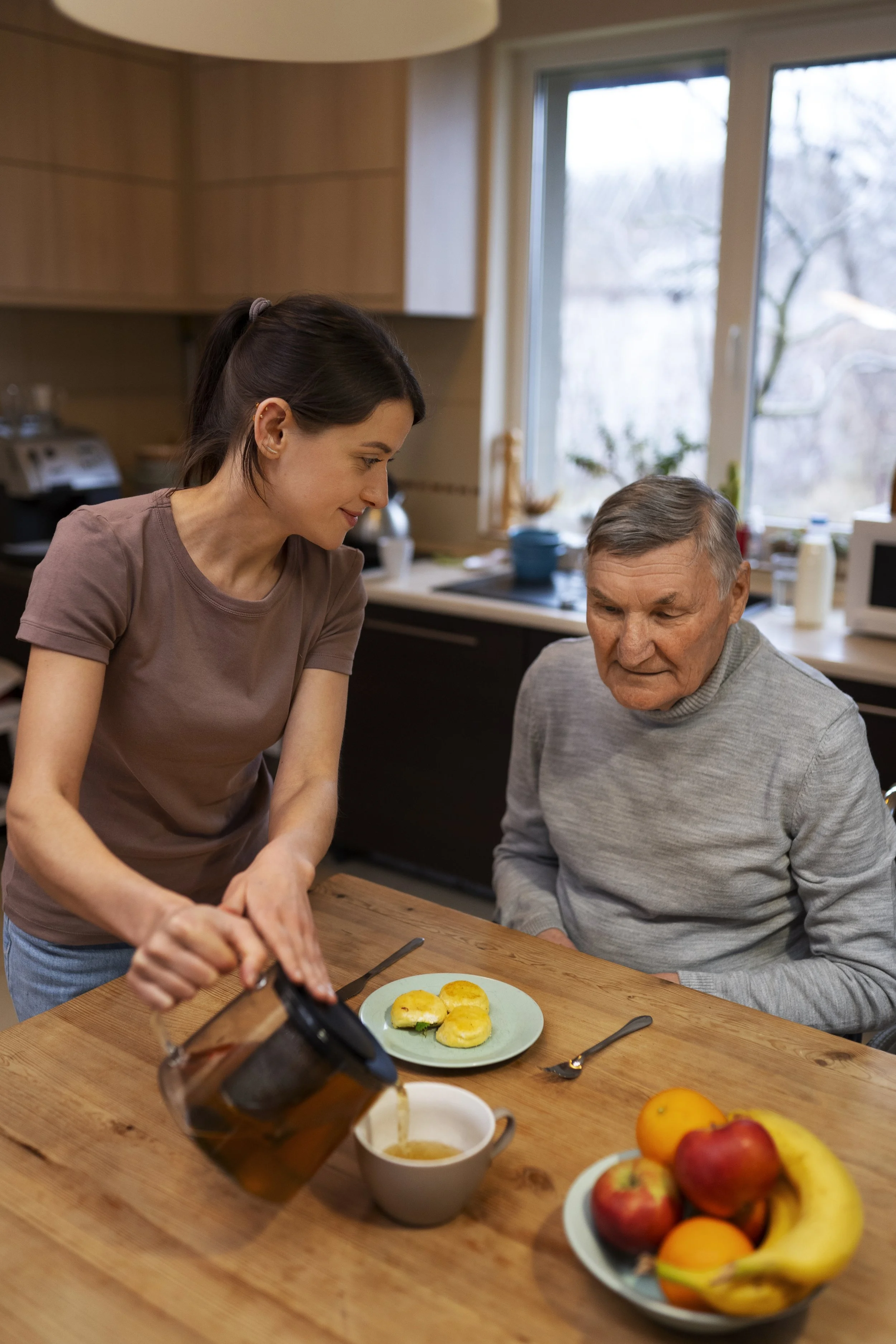 Une jeune femme verse du thé dans une tasse alors qu'un homme âgé regarde, assis à une table en bois avec des assiettes de fruits et de petits pains, dans une cuisine lumineuse.