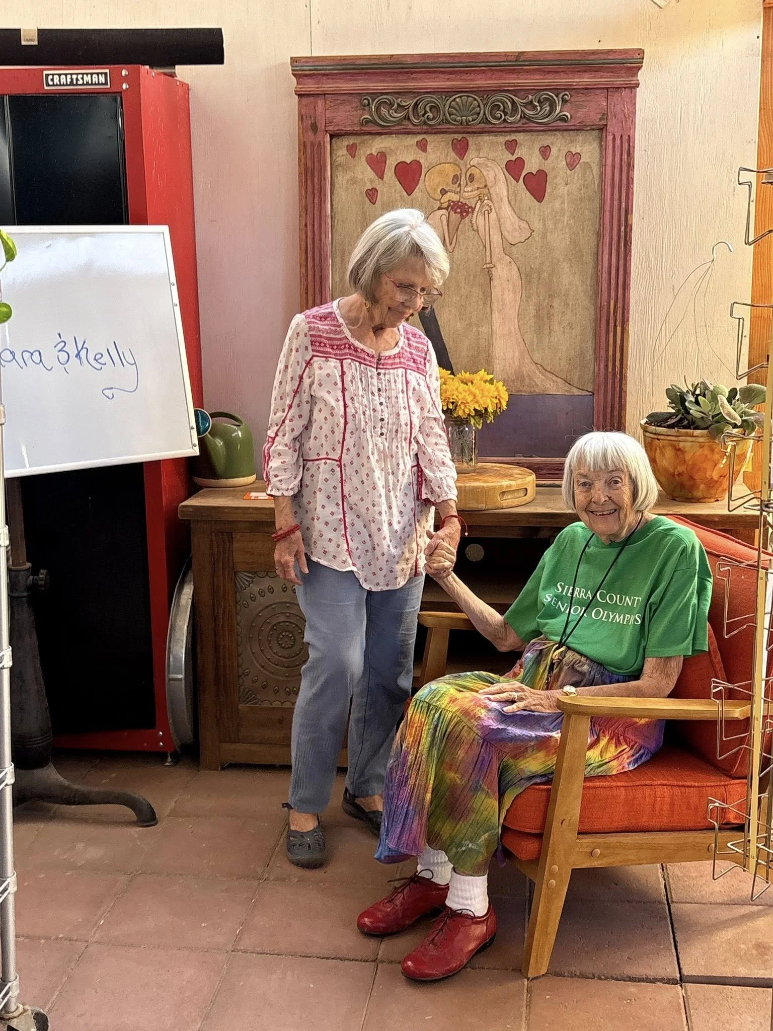 An elderly woman sitting in a wooden armchair, wearing a green event T-shirt and colorful tie-dye pants, holding hands with a woman standing next to her. The elderly woman is smiling. The setting appears to be indoors with decorative art and plants in the background.