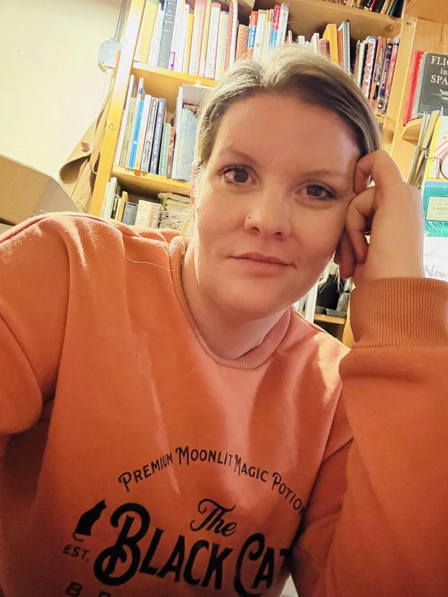 A woman with light brown hair, wearing an orange sweatshirt with black text, sitting in front of a bookshelf filled with books, resting her head on her hand.