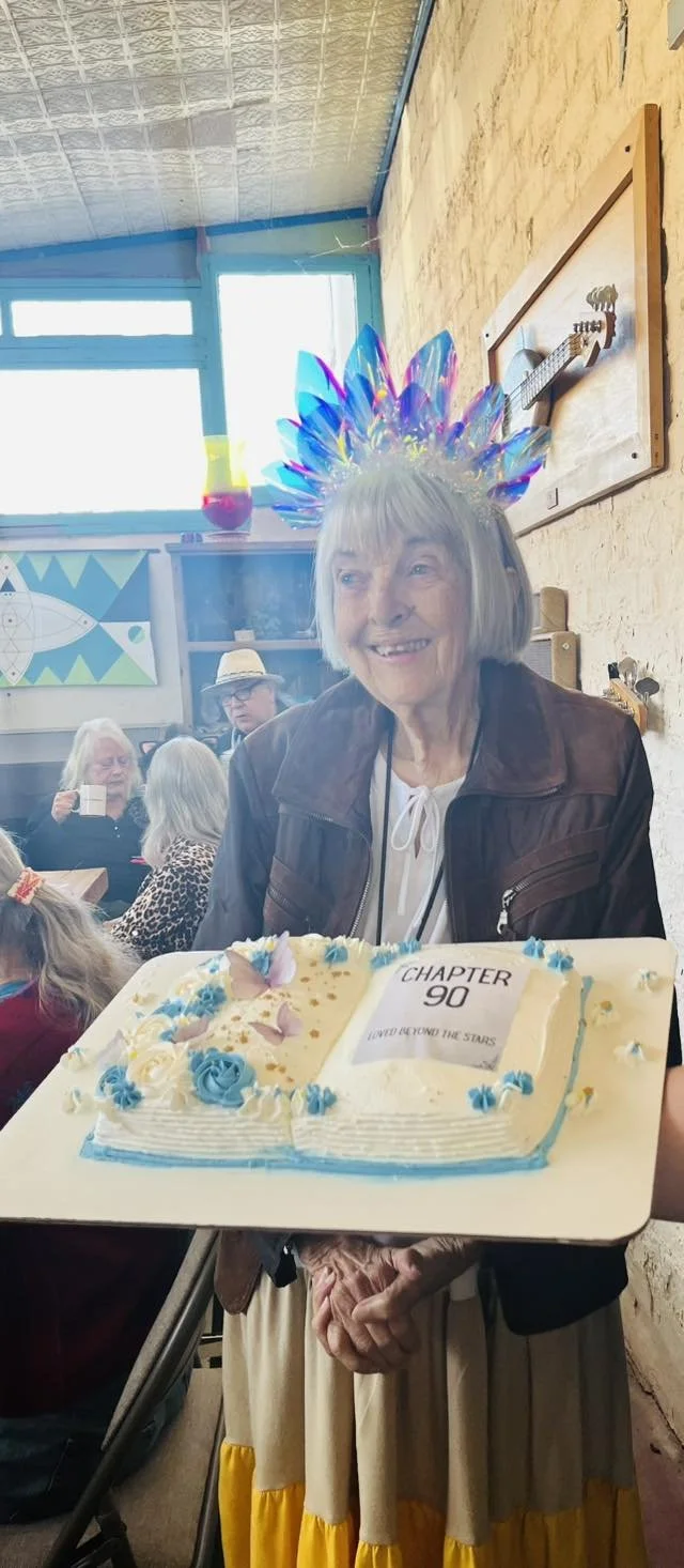 An elderly woman wearing a colorful feathered headband is holding a cake shaped like an open book that says "Chapter 90, Loved to the Stars." She is smiling and there are other people in the background, including an elderly woman with a straw hat and a woman with white hair sipping from a cup. The setting appears to be a celebration or party.