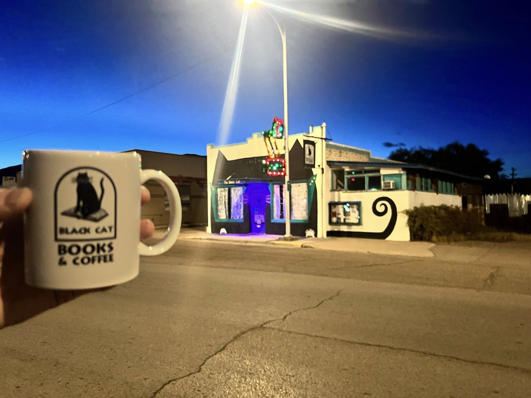 A hand holding a mug with the logo of Black Cat Books & Coffee in front of a street scene at night featuring a colorful, illuminated building with a neon sign and a blue-lit window.