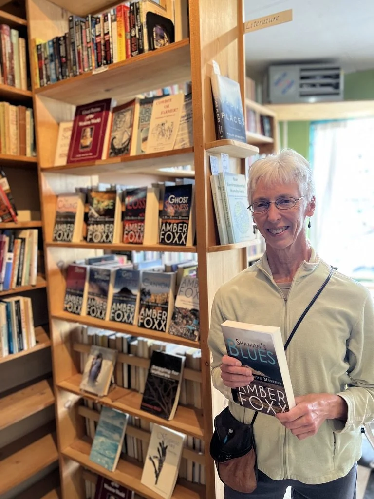 A woman with short white hair and glasses smiling while holding a book titled 'Shaman's Blues' by Amber Foxx in a bookshop. Behind her are shelves filled with various books.