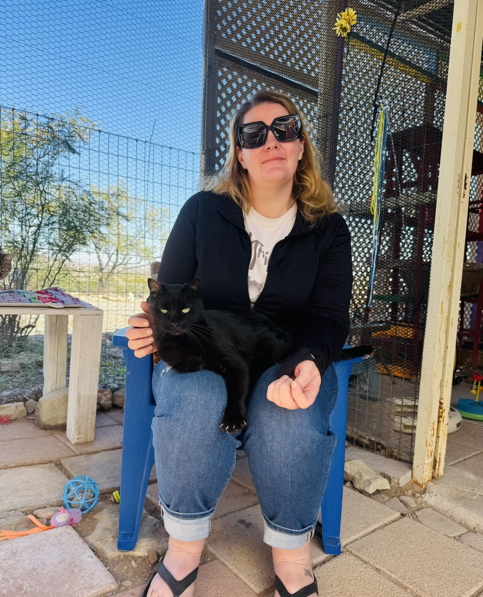 A woman with long, wavy hair wearing black sunglasses, a black jacket, a white t-shirt, and rolled-up jeans sitting outside on a blue plastic chair, holding a black cat with green eyes. The background shows a fenced area with outdoor toys, a small table with colorful items, and a clear blue sky.