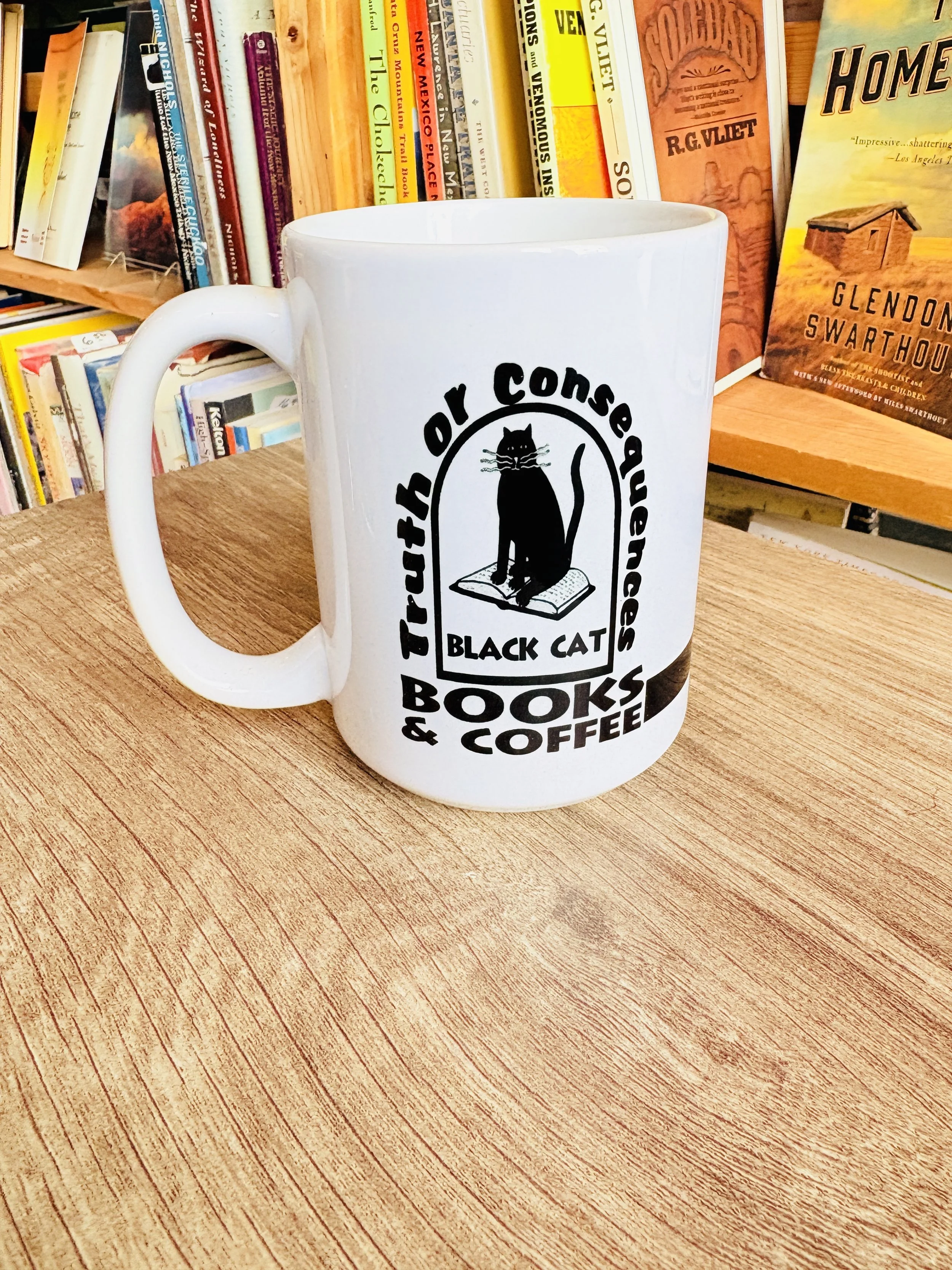 White mug with black text and a graphic of a black cat sitting on a book, with the text 'Truth or Consequences Black Cat Books & Coffee' on a wooden table in a bookstore.
