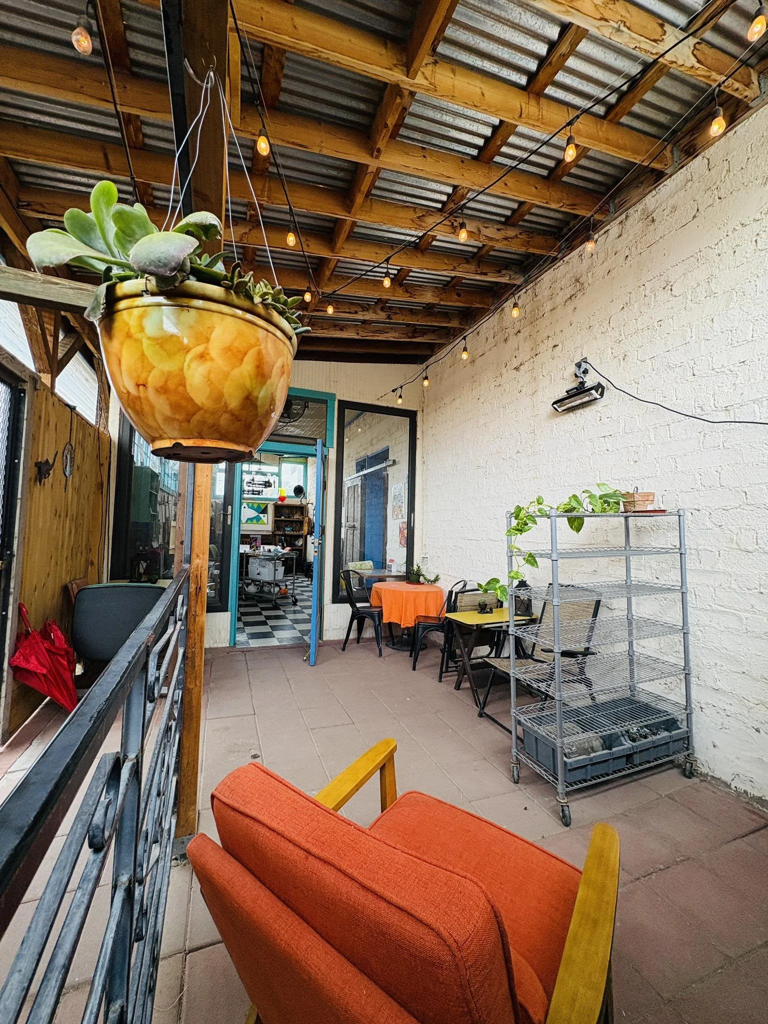Outdoor patio with orange cushioned chair, hanging potted plant, string lights, white brick wall, metal shelving unit with plants, and table with orange tablecloth.