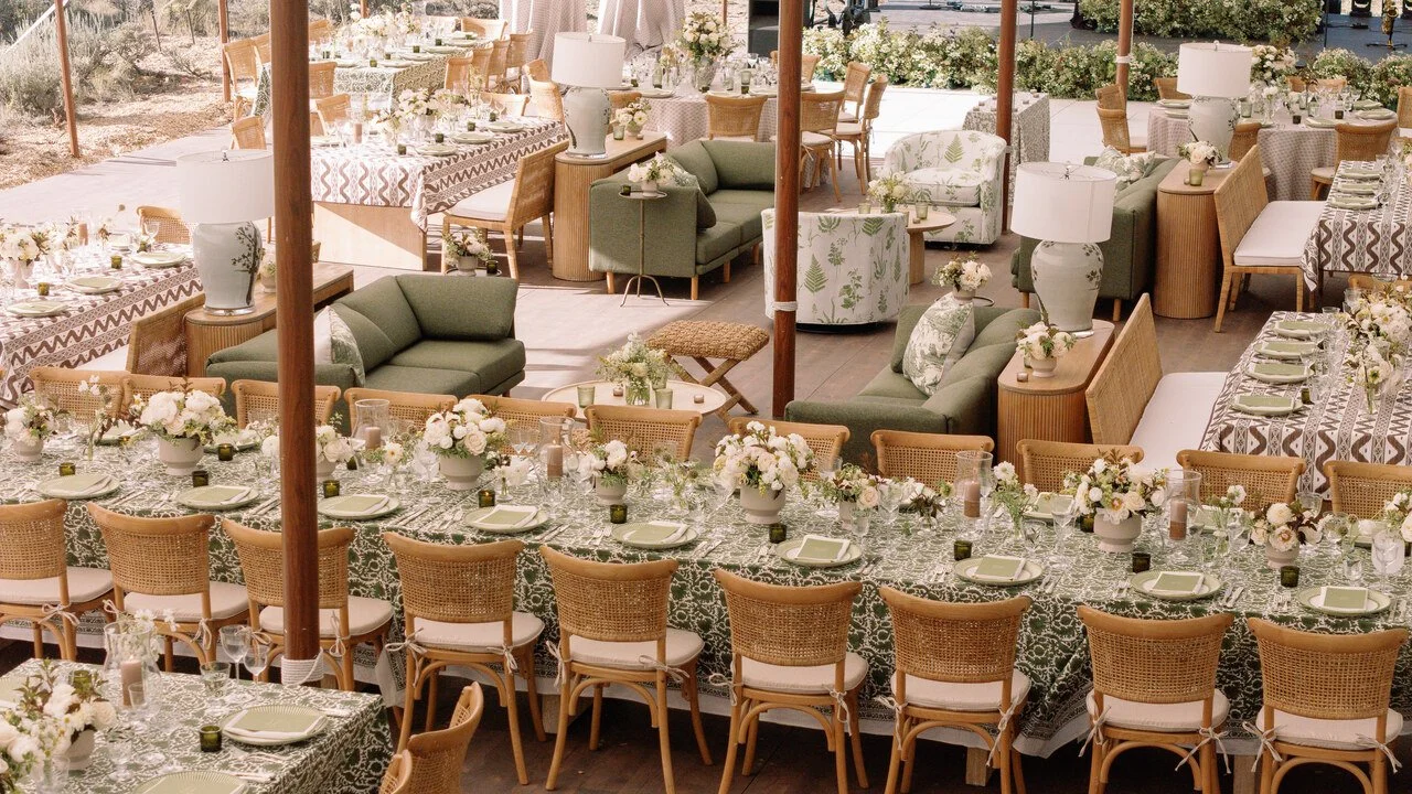 Elegant outdoor dining setup with long tables covered in patterned tablecloths, adorned with floral centerpieces, surrounded by wicker chairs. There is a cozy lounge area with green sofas, armchairs, lamps, and small side tables, set under a tent with natural light and decorative floral arrangements.
