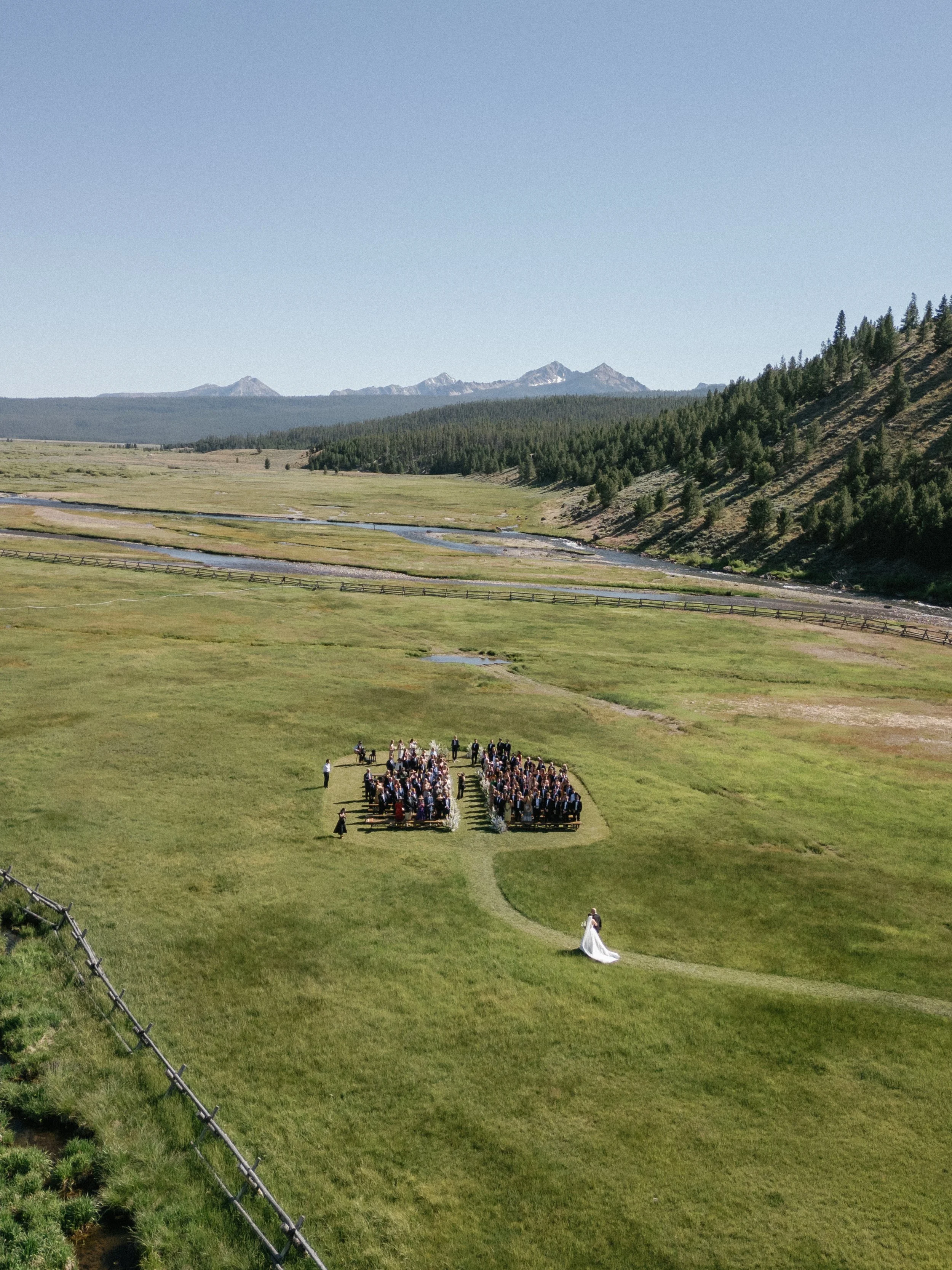 A wedding ceremony taking place outdoors on a grassy field, with a bride in a white wedding dress and a group of guests seated on benches, mountains and trees visible in the background.