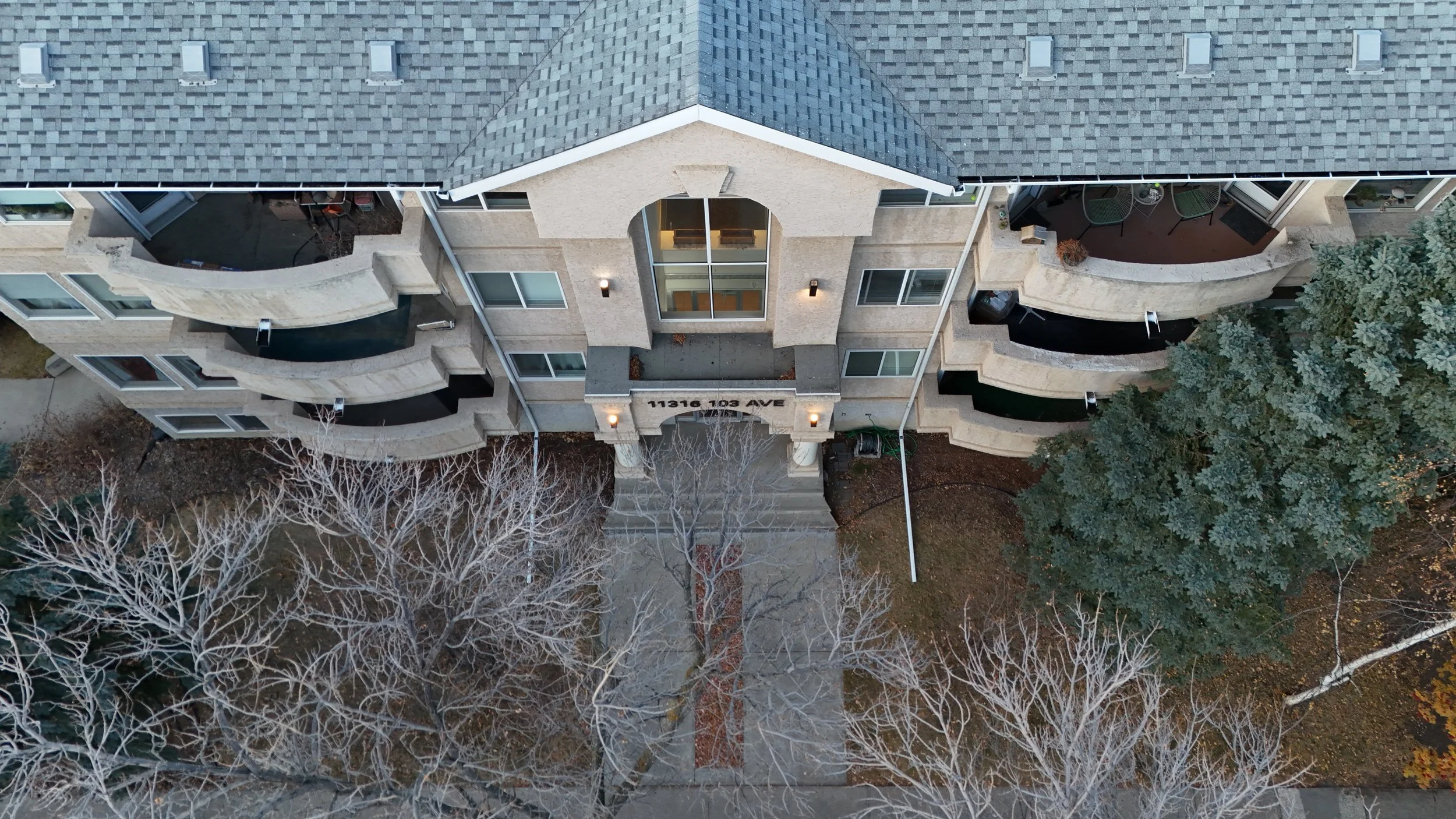 Aerial view of a multi-story apartment building with balconies and a central entrance, surrounded by leafless trees and some evergreen trees.