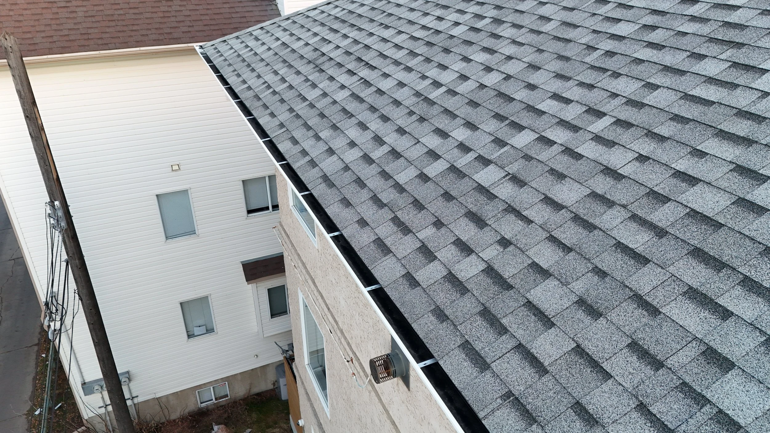 A bird's eye view of rooftops, showing a gray shingled roof and a white siding building with windows.