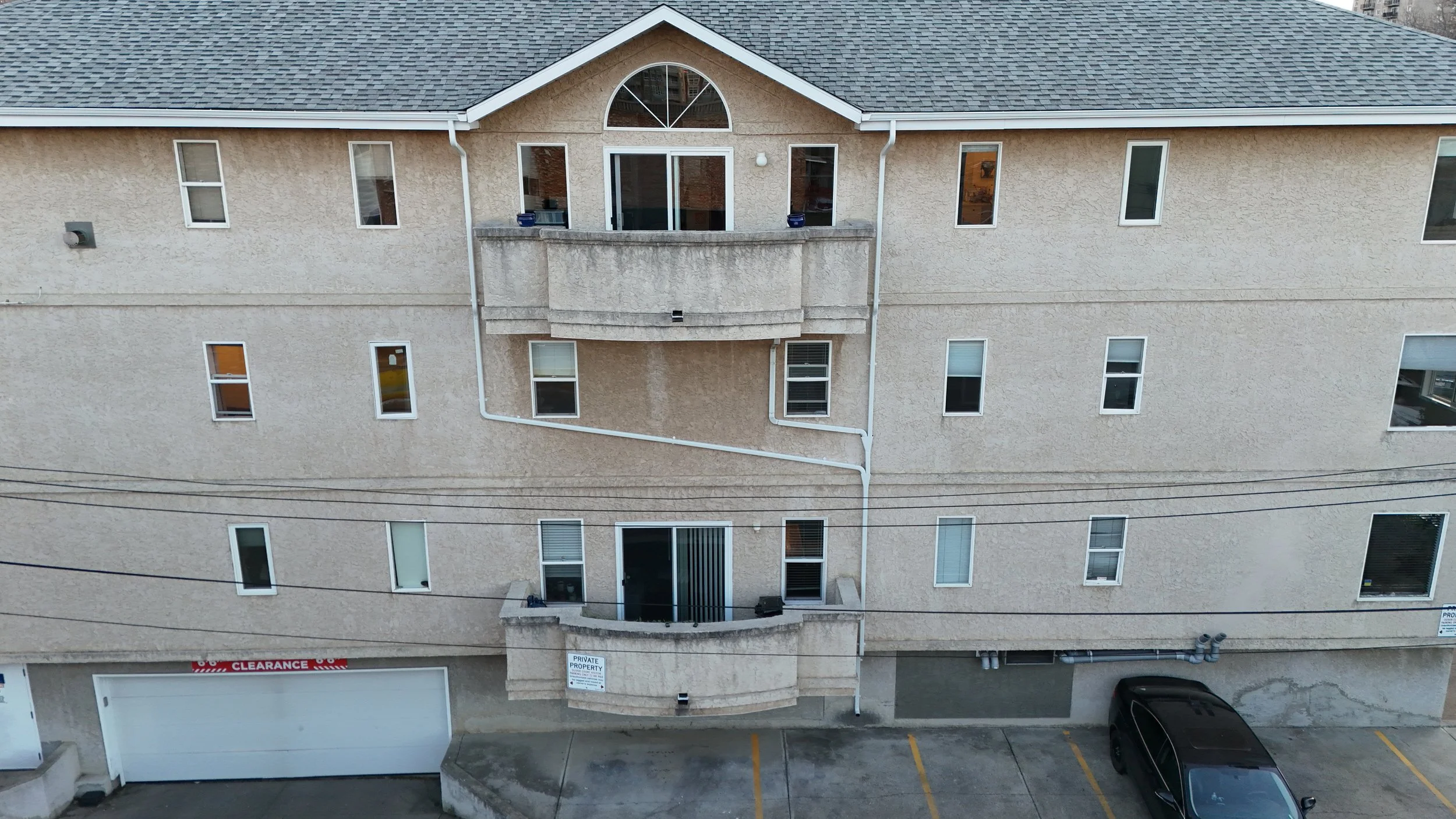 Three-story beige apartment building with multiple windows, balconies, and a garage, parked car, utility pipes, and electrical wires in the parking lot.