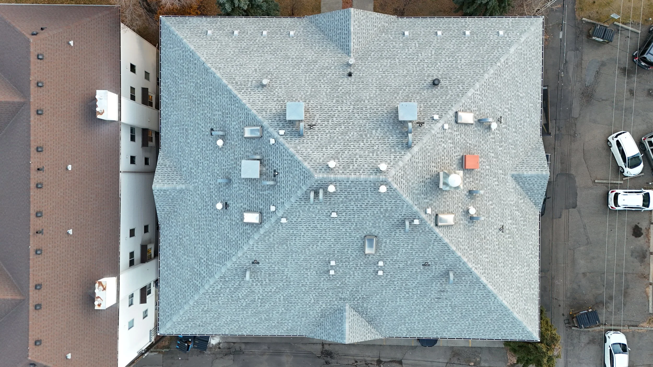 Top-down aerial view of a building with a gray shingle roof and a brown and white adjacent building, surrounded by parked cars and parking lot.