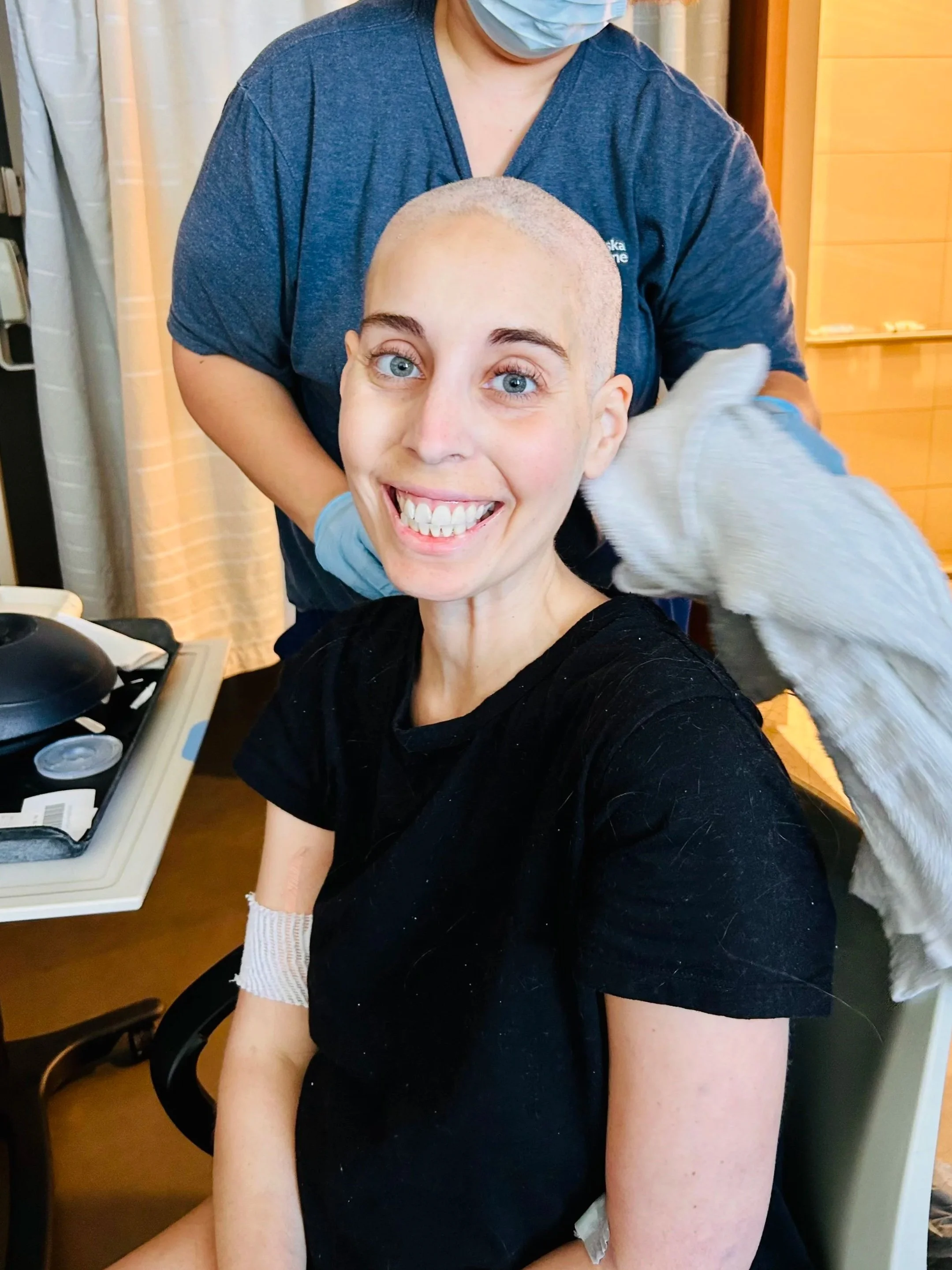 A woman with a shaved head smiling at the camera while a nurse or medical professional assists her, possibly in a hospital or clinic setting.