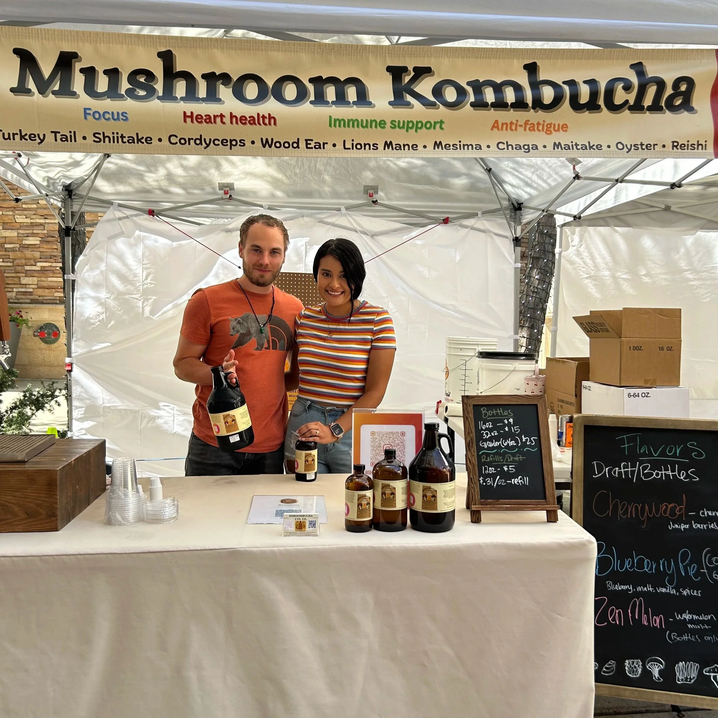 Two people standing behind a table at a booth, a man holding a bottle of syrup, at a market or fair. A sign above reads "Mushroom Kombucha" with benefits listed below, and blackboards with menu items and prices are on the table.
