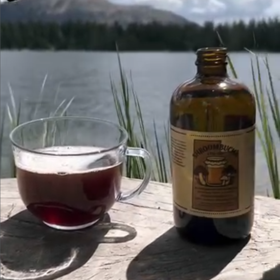 A glass mug filled with a dark beverage and an amber bottle with a yellow label next to it, both placed on a wooden surface by a lakeside with trees and mountains in the background.