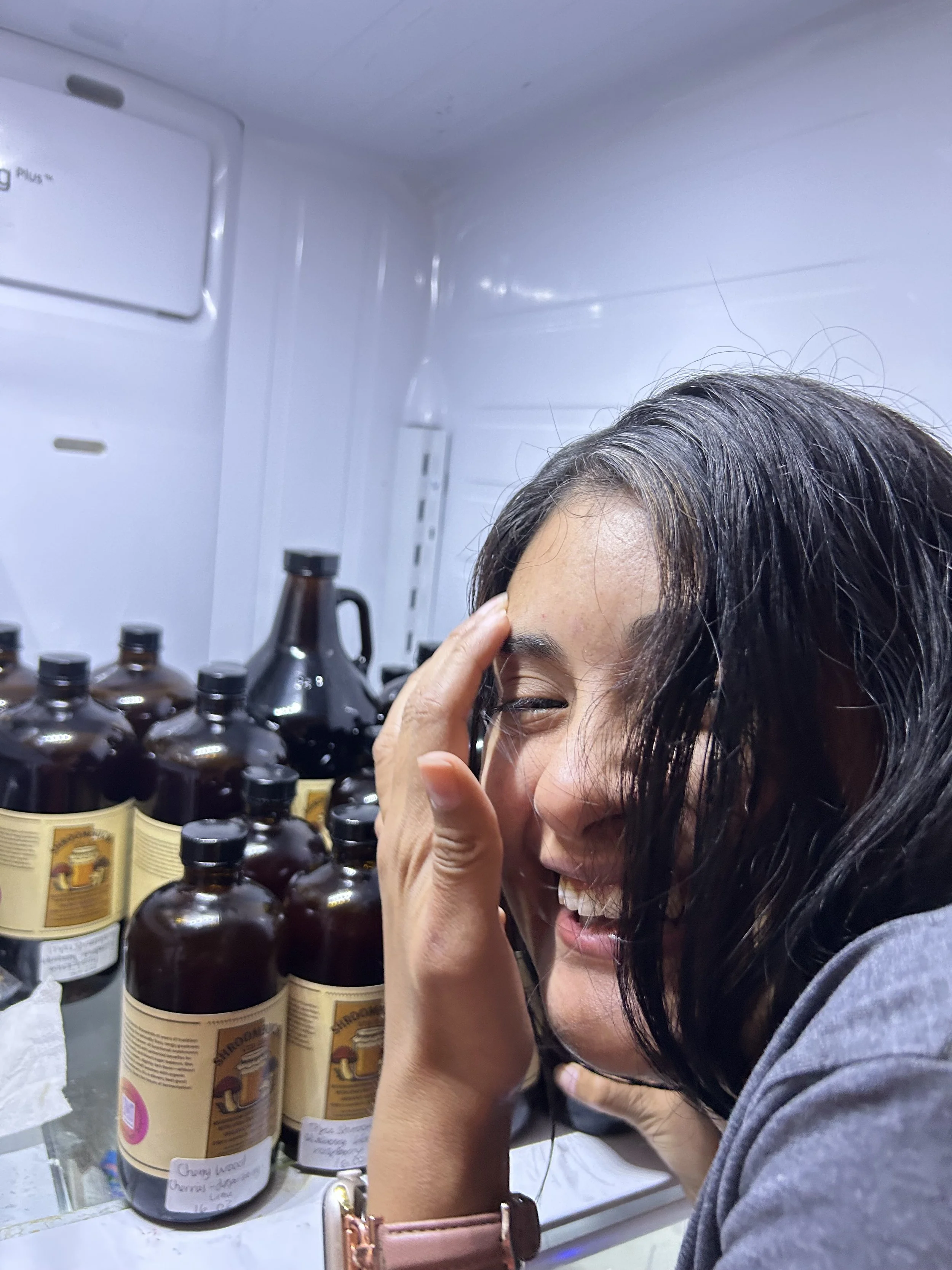 A woman with dark hair smiling and touching her face in a refrigerator with brown bottles and labels inside.