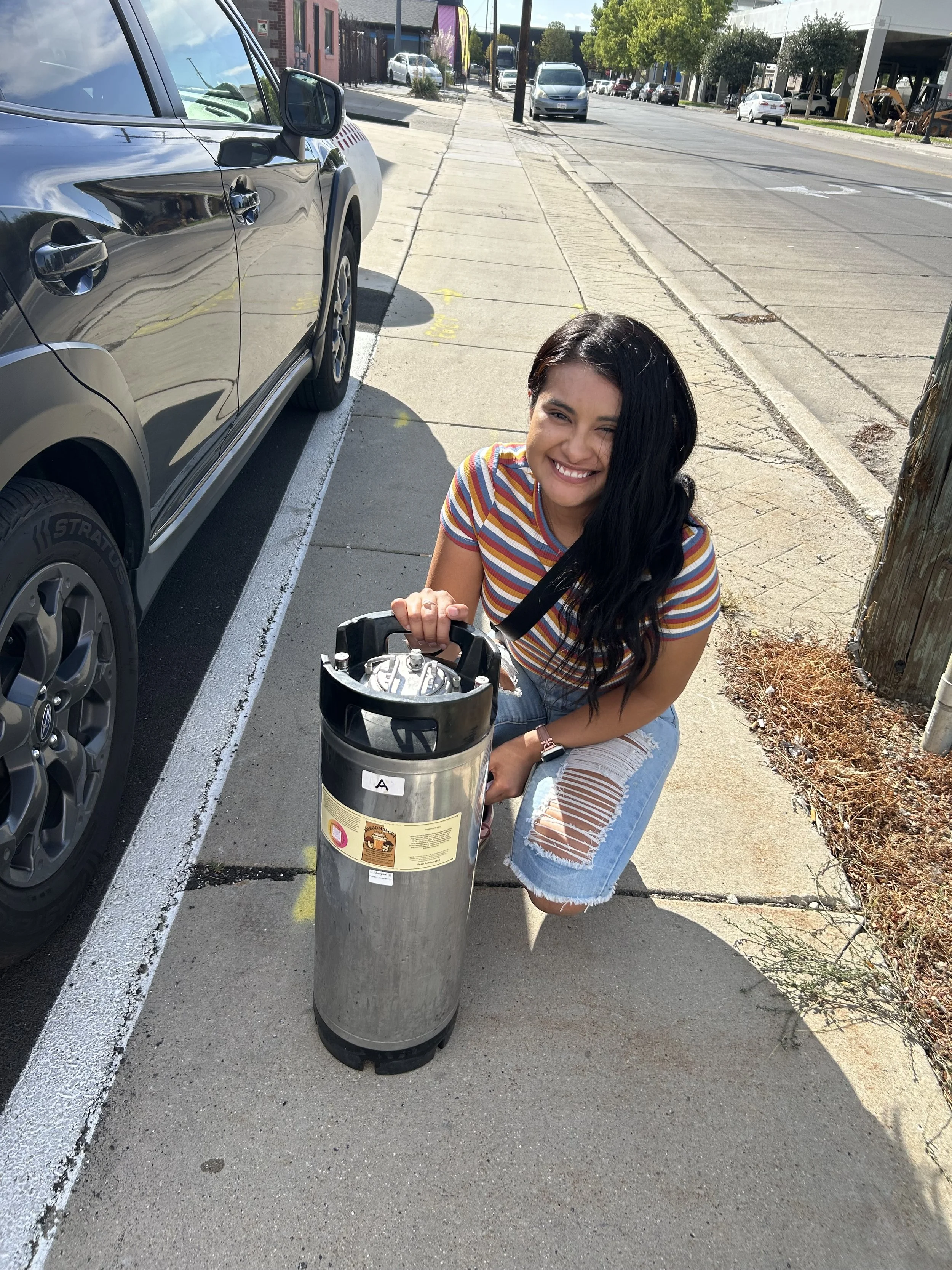 A young woman with long dark hair, wearing a striped t-shirt and ripped jeans, smiling while bending down on a sidewalk next to a trash can on a city street.