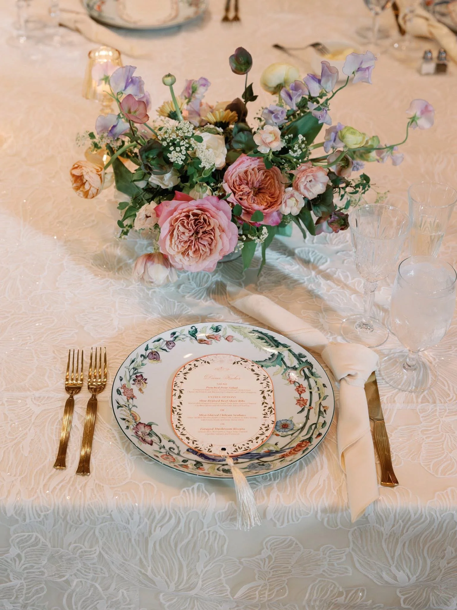 Elegant table setting with a floral centerpiece, a decorative dinner plate, gold cutlery, clear wine glasses, and a white napkin tied around the utensils on a cream-colored tablecloth with a floral pattern.