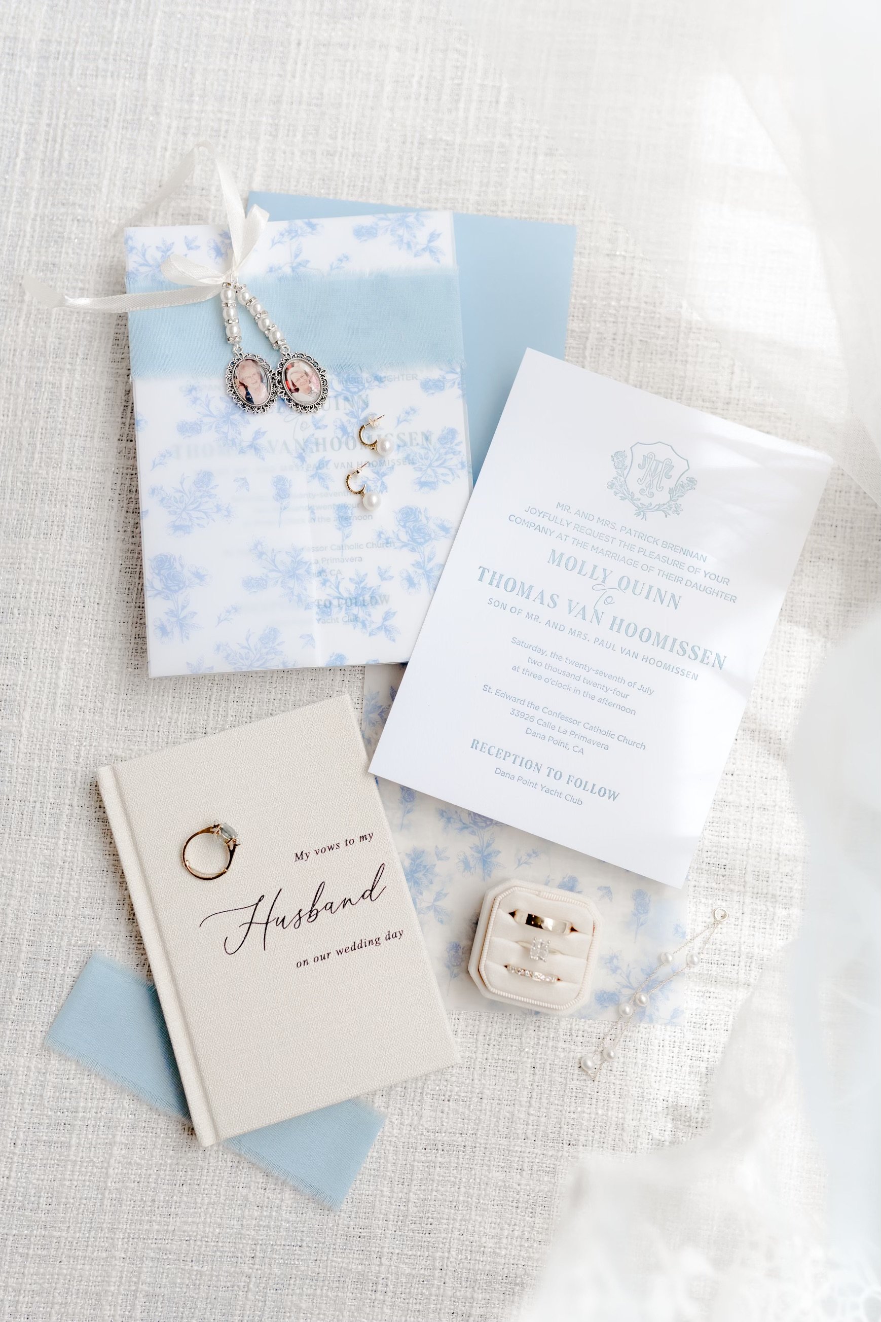 Wedding invitation, jewelry, and vow book on a white textured surface with sheer curtain.